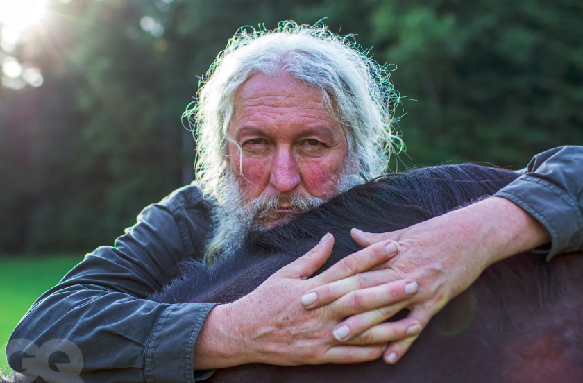 An elderly man with long white hair and beard hugging a black horse outdoors with trees in the background.