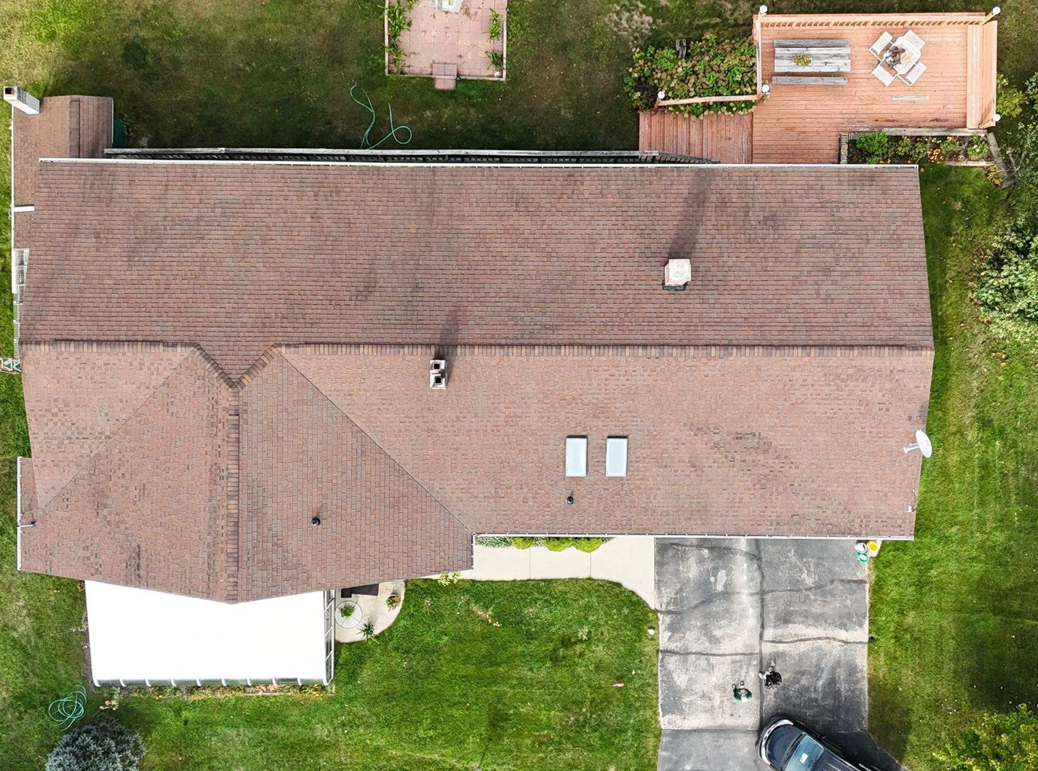 Aerial view of a house with a brown shingled roof, a driveway with a car parked at the end, a small backyard, a wooden deck with outdoor furniture, and a garden with plants and a pathway.