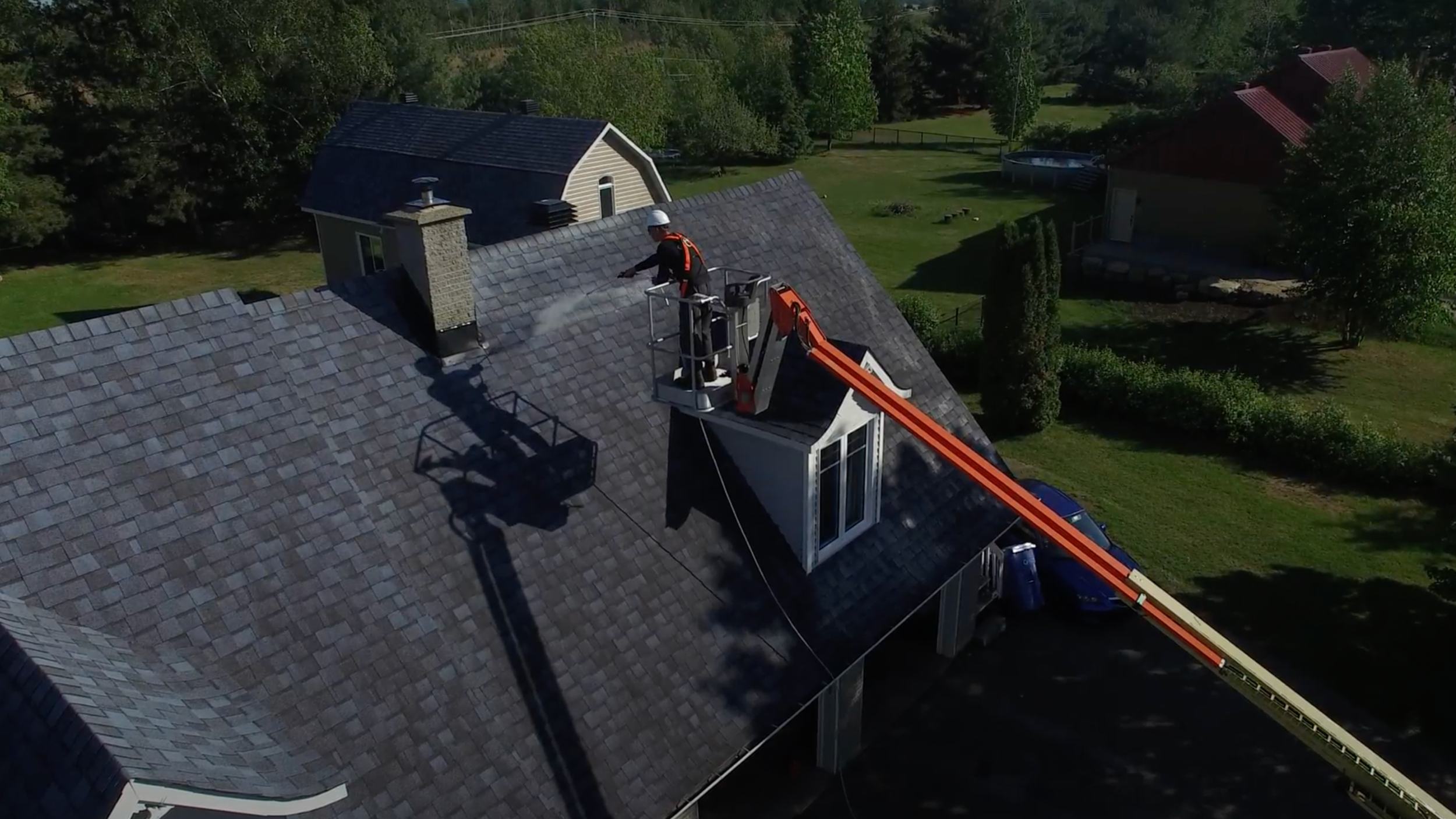 A worker on a lift cleaning a roof with a pressure washer among area trees and neighboring houses.