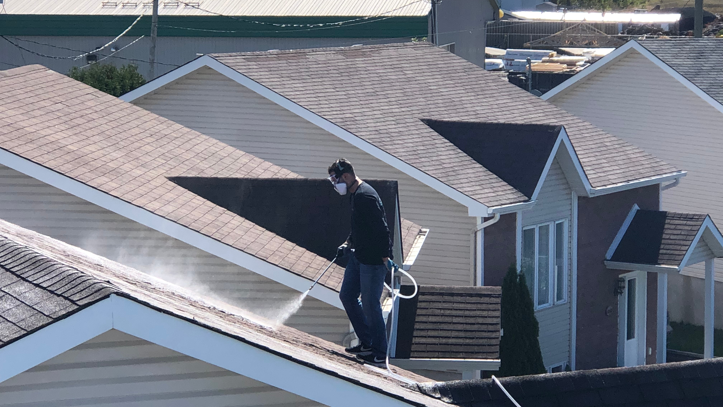 A man wearing a face mask, black jacket, and jeans stands on a house roof, spraying a cleaning solution on the shingles with a spray wand.