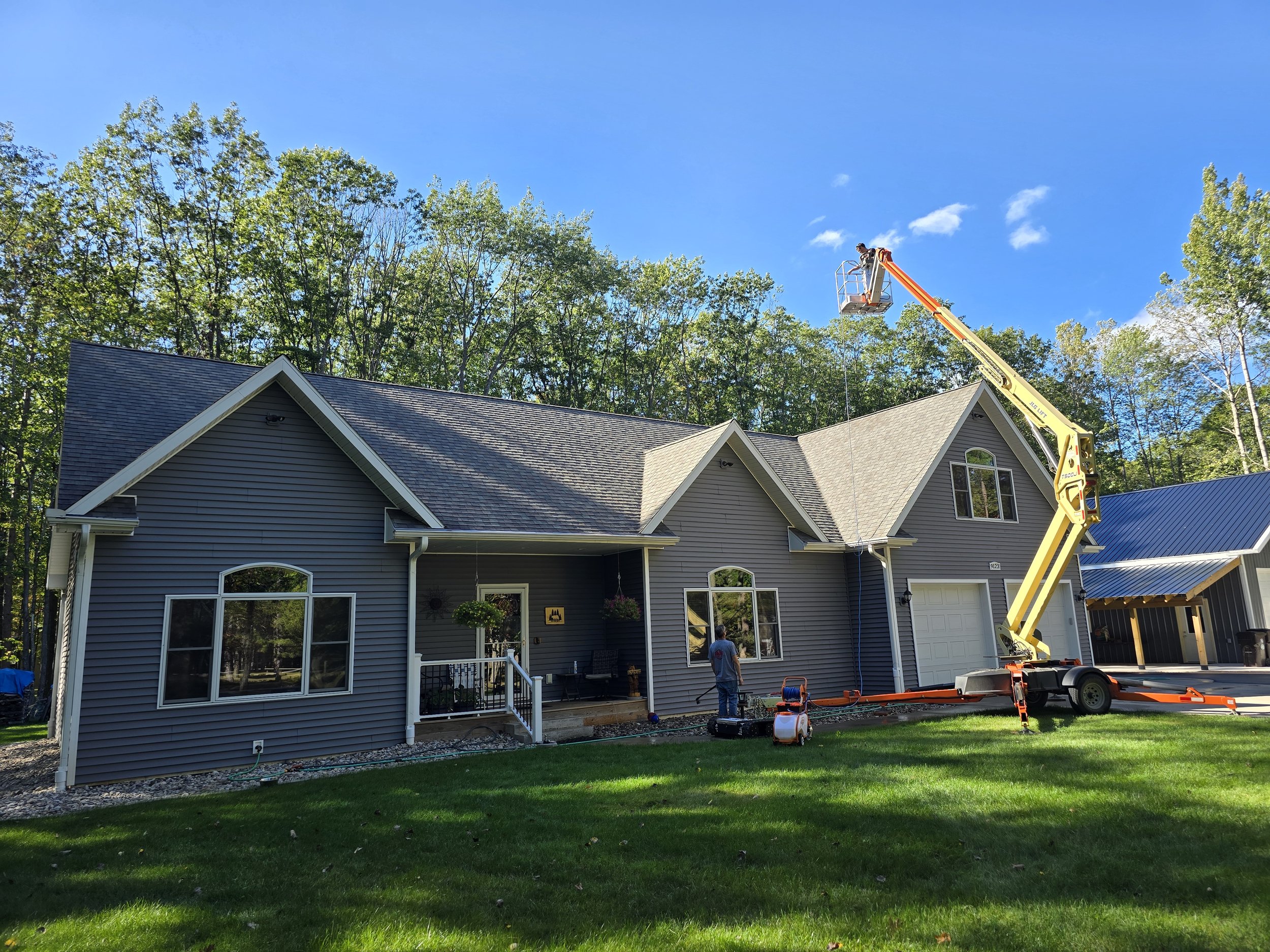 A house with grey siding and a garage being worked on using a cherry picker lift with a worker in the basket, set against a background of trees and a blue sky.