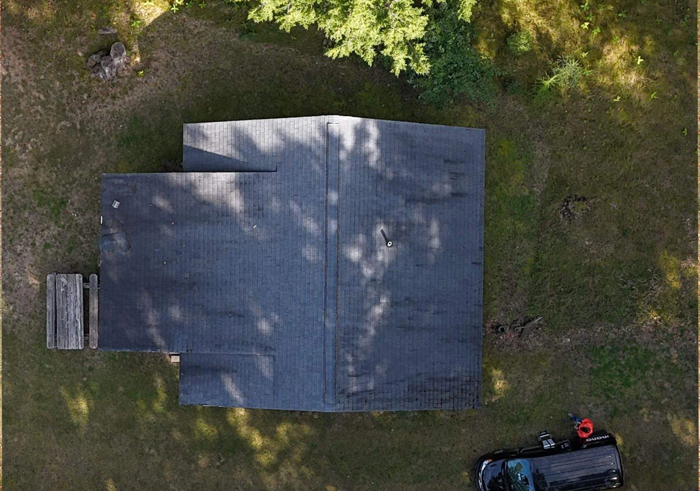 An aerial view of a building with a gray shingle roof, a small wooden bench on the left, a vehicle partially visible at the bottom right, and trees casting shadows over the roof and surrounding grassy area.