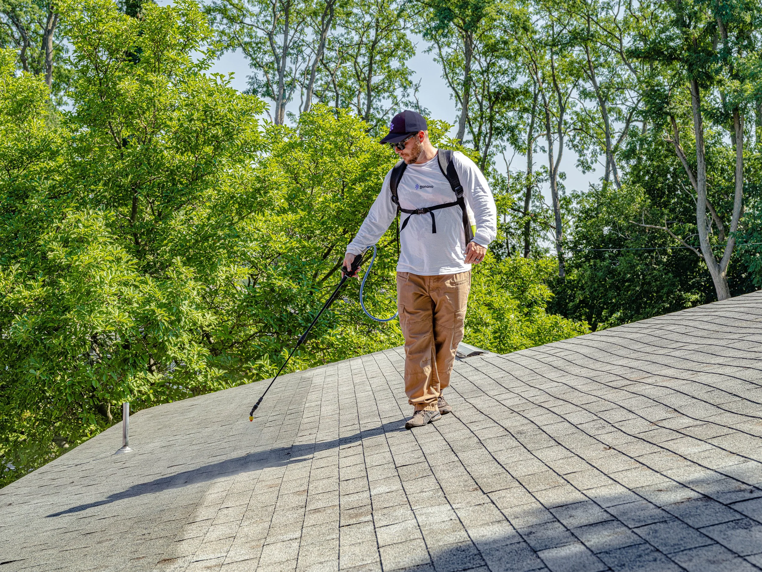 A man walks on a residential roof while using a roof inspection tool, surrounded by green trees.