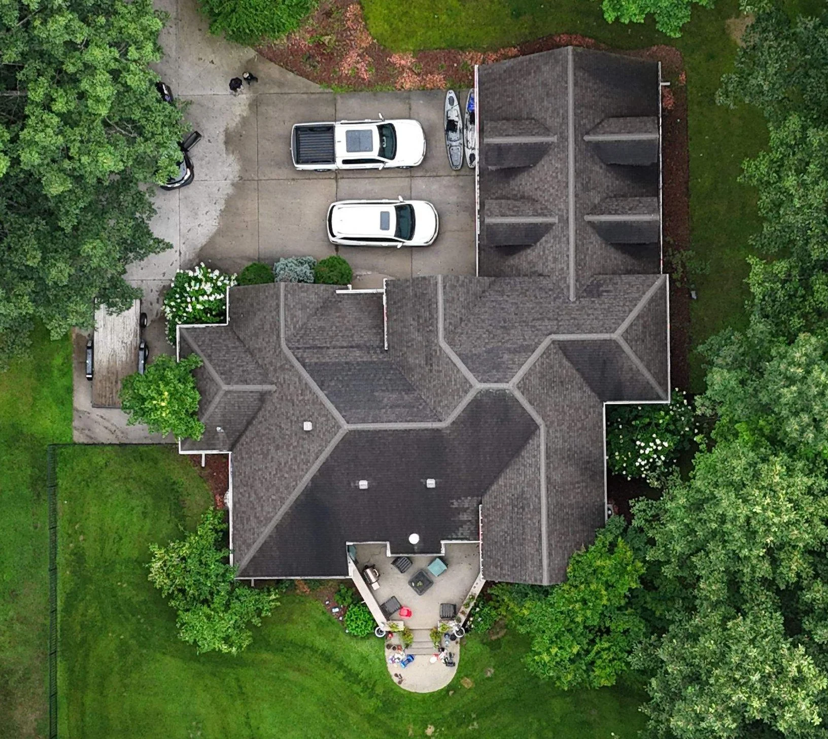 Aerial view of a house with a gray roof, surrounded by green trees and a landscaped yard. There is a circular patio with outdoor furniture at the back of the house. The driveway has three white vehicles parked and is bordered by more trees and shrubs.