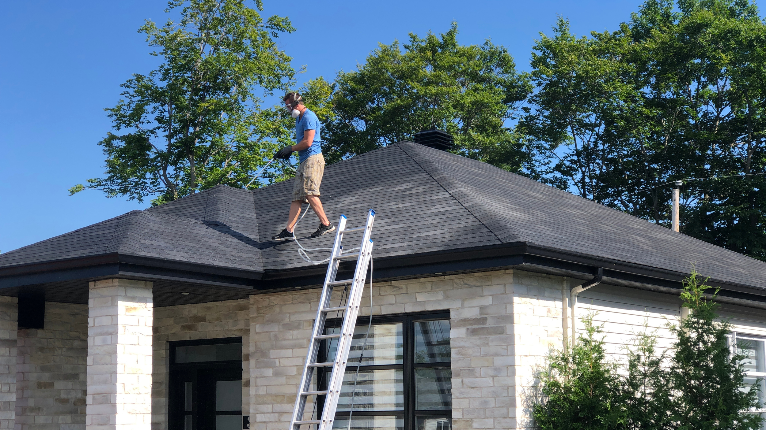 A person cleaning the roof of a house with a sprayer, standing on the roof with a ladder leaning against the house.