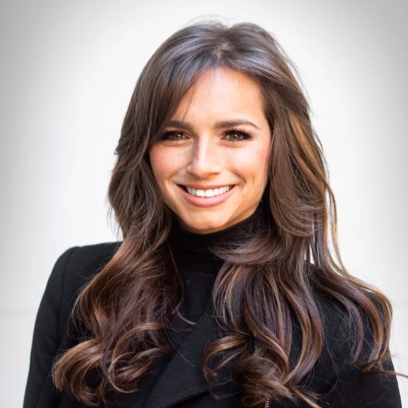 A woman with long wavy brown hair smiling, wearing a black blazer, against a plain light background.