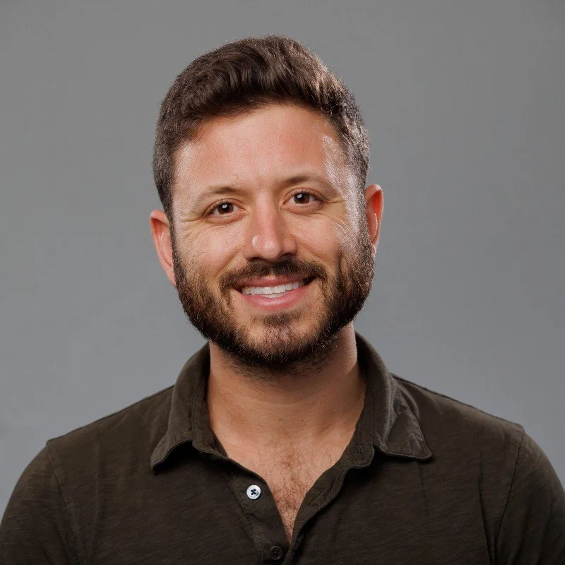 A smiling man with a beard, wearing a dark button-up shirt, against a plain gray background.