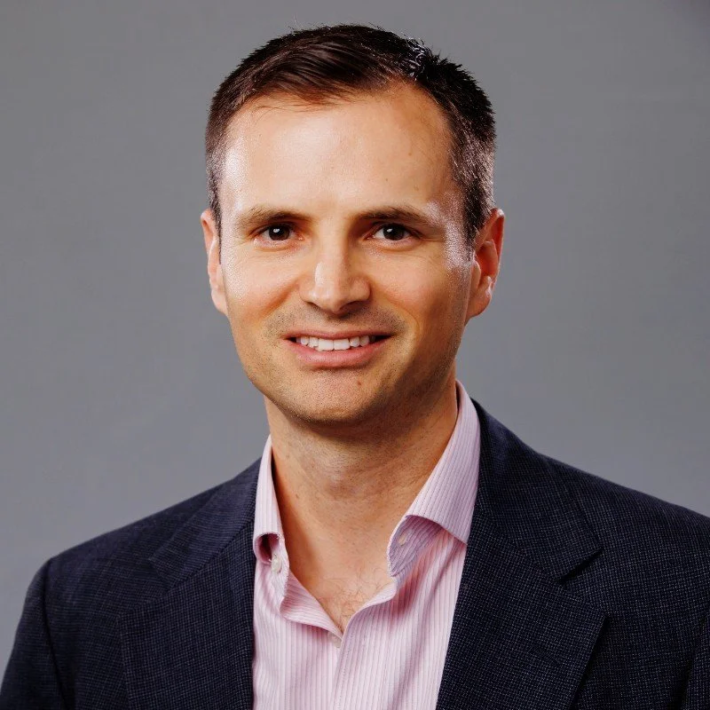 Headshot of a smiling man with short dark hair wearing a pink collared shirt and a dark blazer, against a gray background.