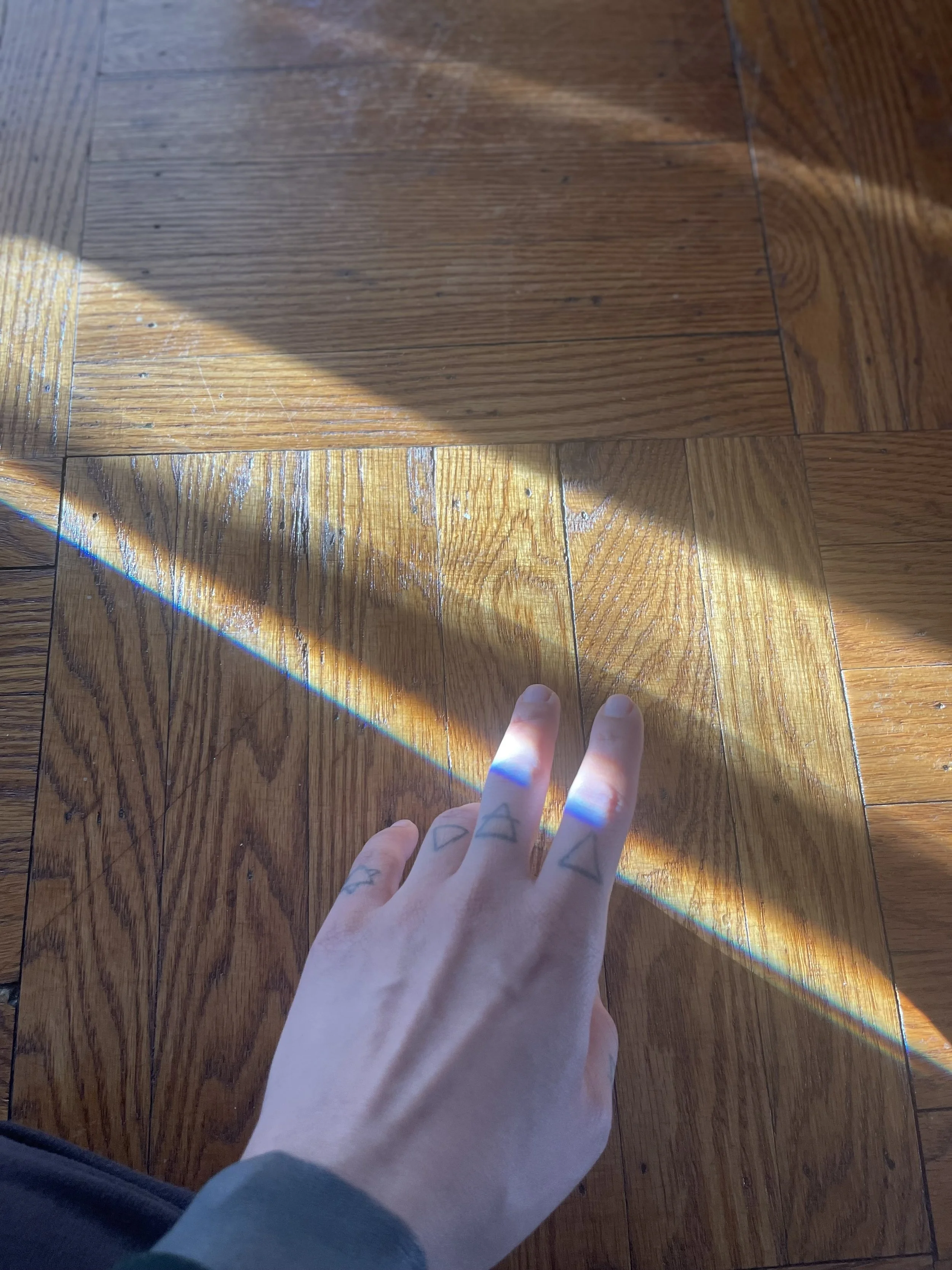A person's hand with geometric tattoos is pointing at a wooden floor, and a rainbow-colored light beam is reflected on the floor.