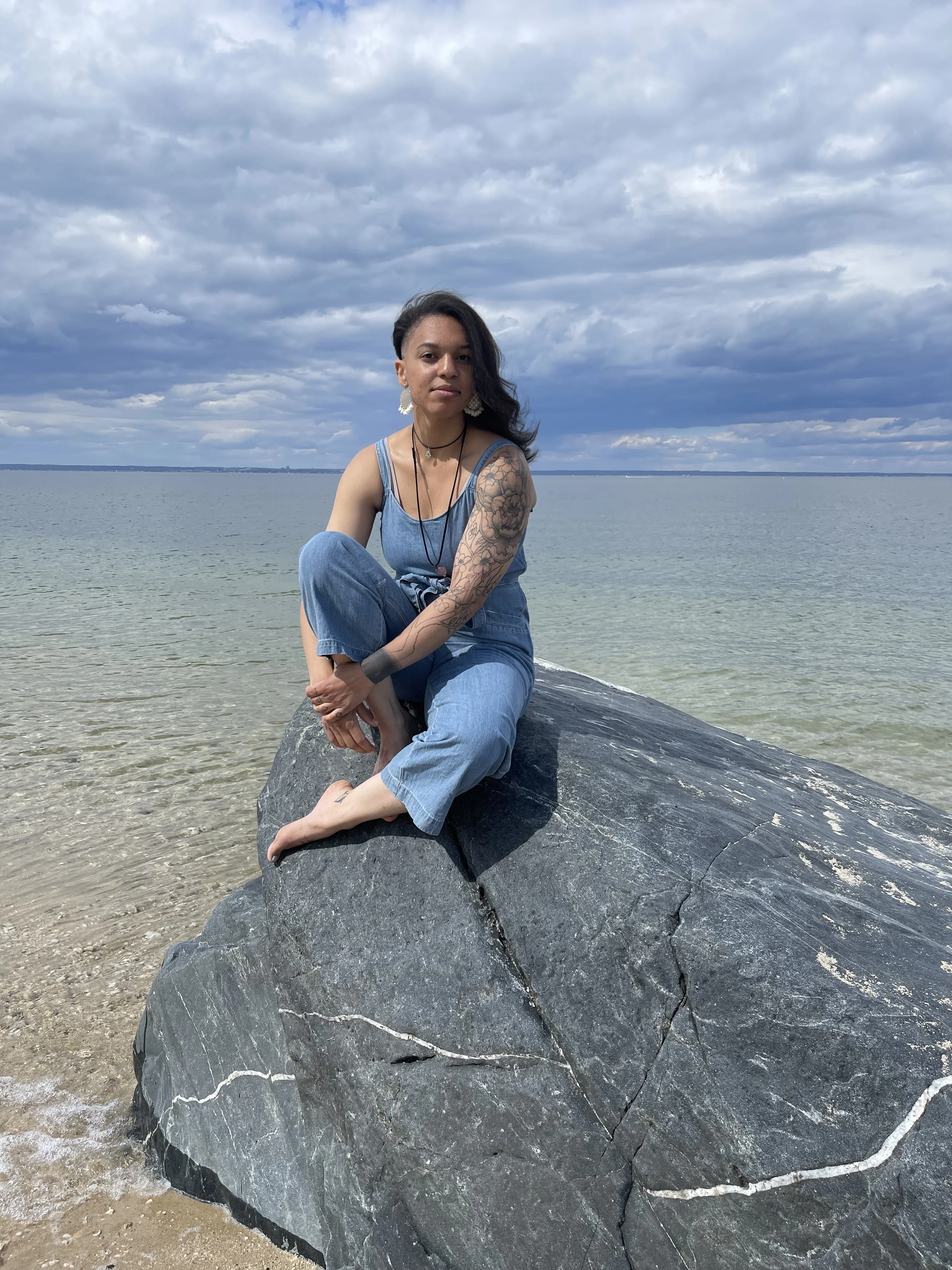 A woman with tattoos sitting on a large black rock at the beach, ocean and cloudy sky in the background.