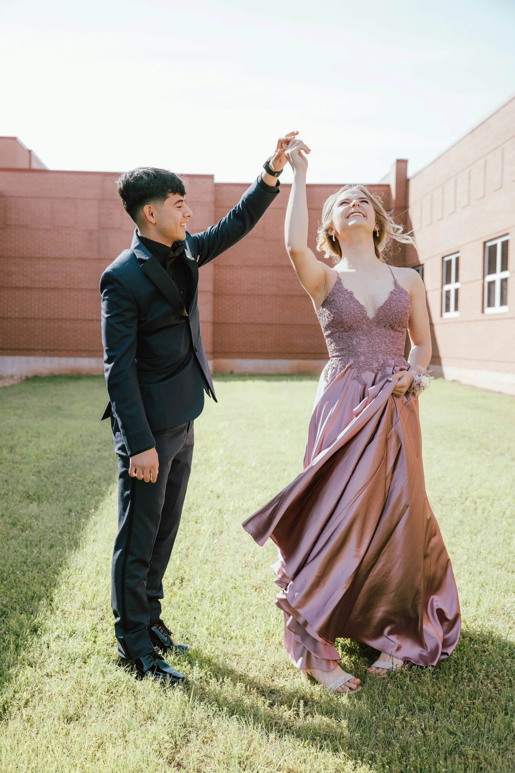 A young couple dancing outdoors on a sunny day, with the man in a dark suit and the woman in a purple gown, in front of a brick building.