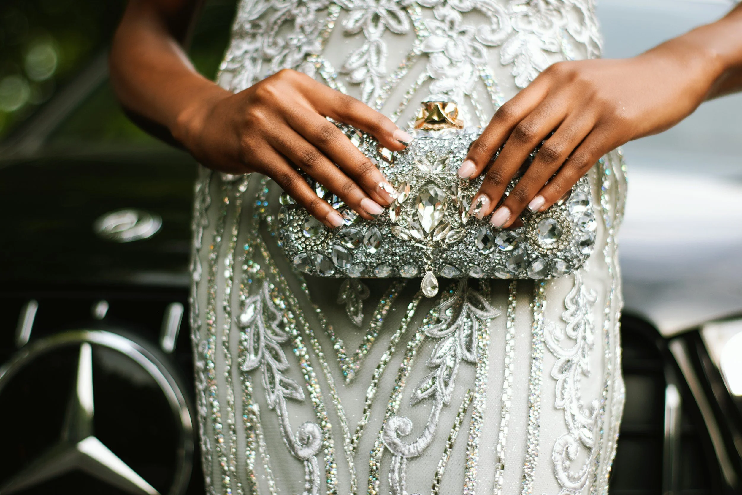 A woman's hands holding a large, ornate, diamond-encrusted clutch purse, with her wearing a detailed, beaded, and sequined dress with a floral pattern.