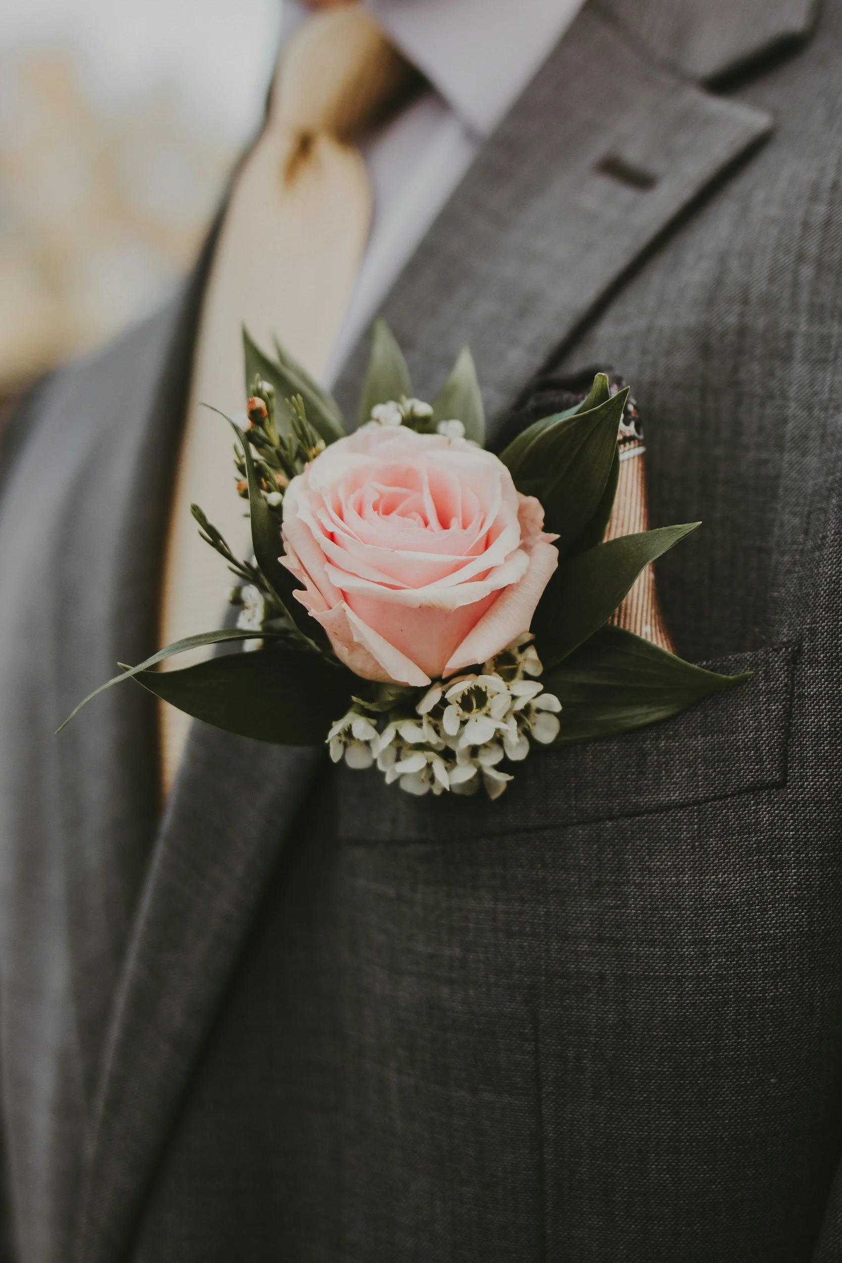 Close-up of a pink rose boutonniere on a gray suit jacket, with greenery and small white flowers.