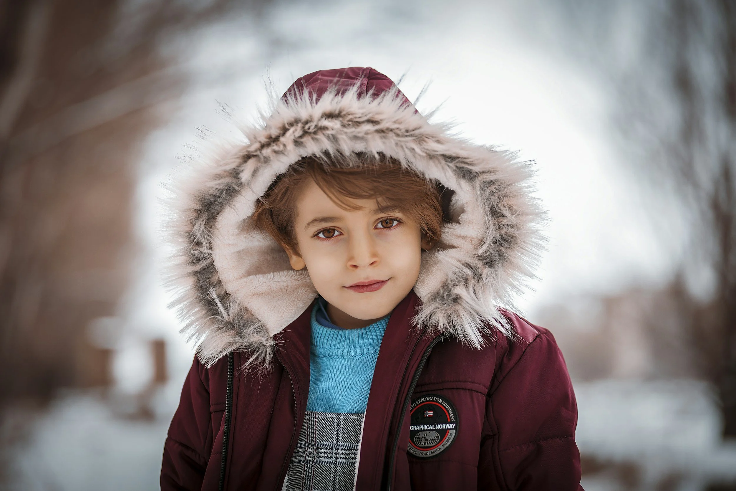 A young boy wearing a maroon winter coat with a fur-lined hood and a blue sweater, standing outdoors in a snowy environment.