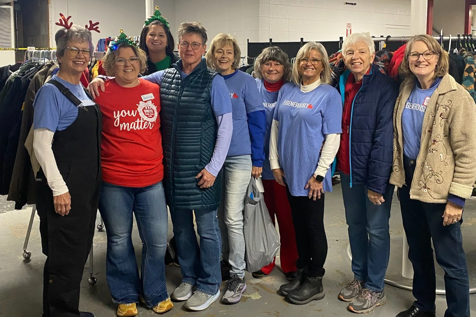 Group of women standing in a line, smiling, at a clothing donation event with racks of clothes in the background. Some wear blue charity shirts, one wears a red shirt that says 'you matter,' and two women wear festive holiday accessories.