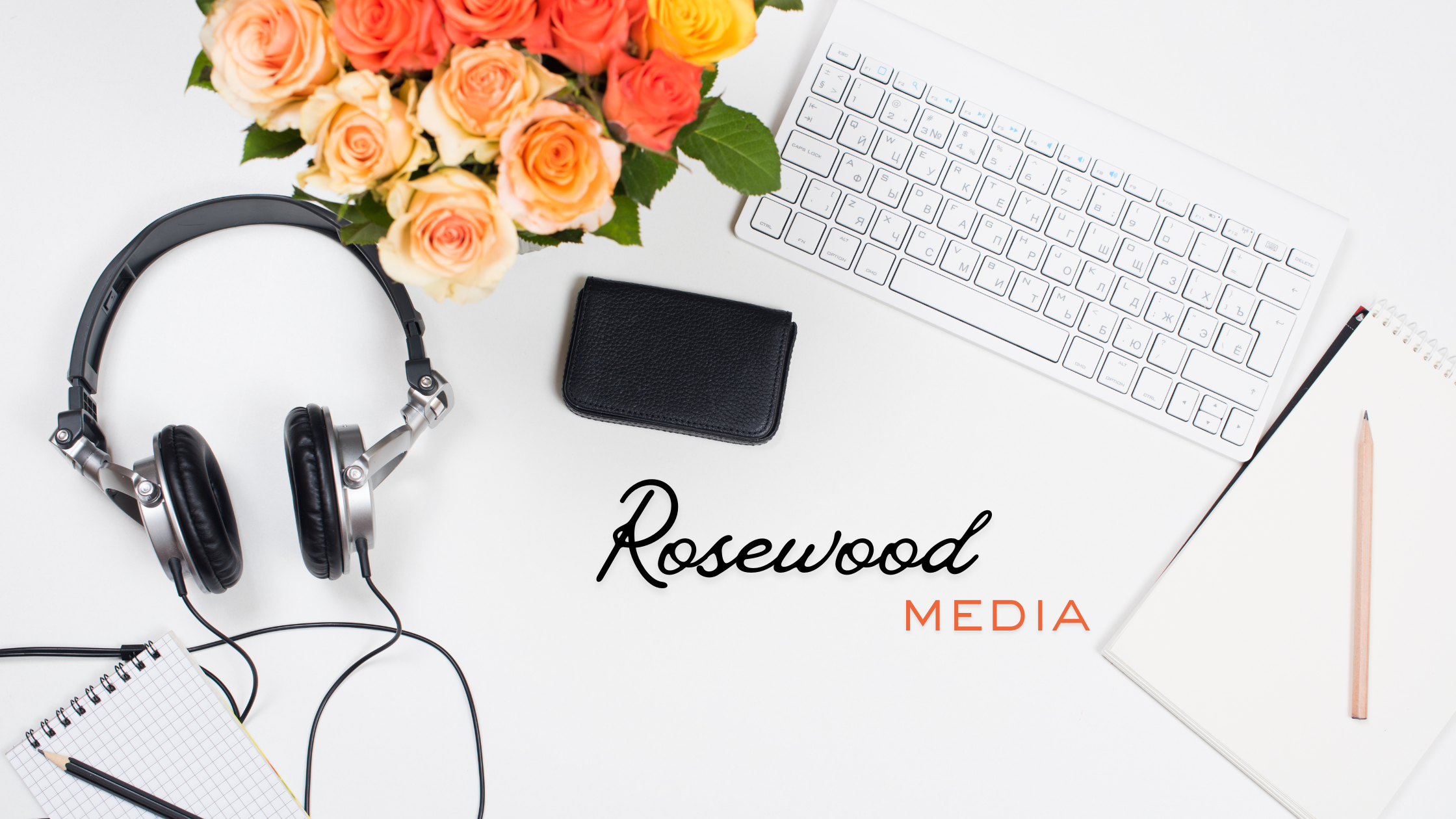 A white desk with a bouquet of orange and pink roses, black and silver headphones, a black wallet, a white wireless keyboard, a white notepad, a pencil, and a small spiral notebook. The text "Rosewood MEDIA" is in black and orange.