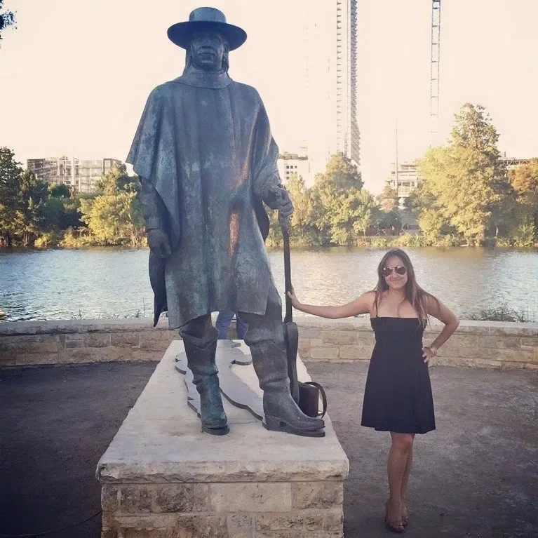 Woman in black dress and sunglasses standing next to a large sculpture of a cowboy holding a lasso and a saddle by a waterfront