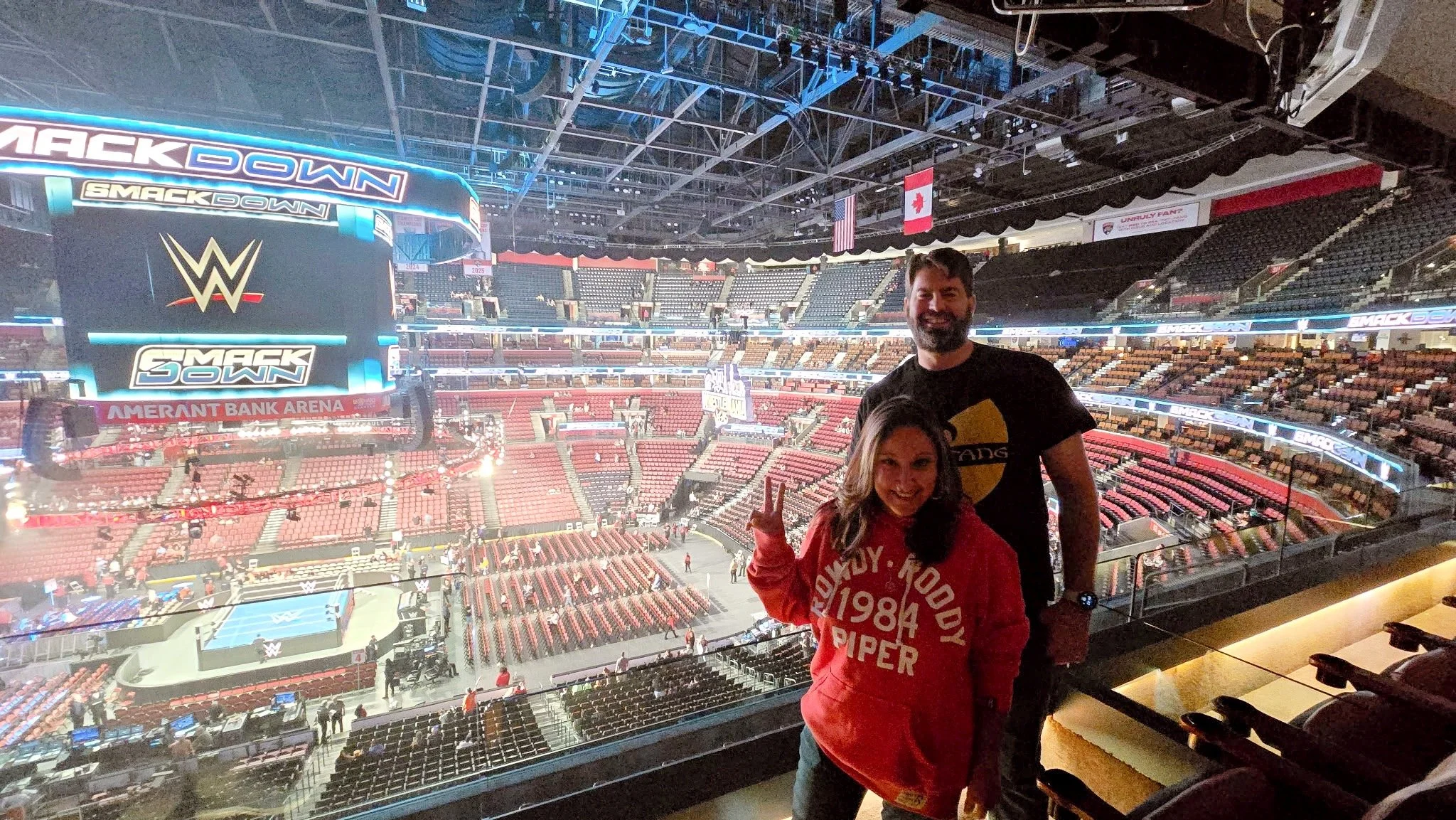 Two people standing inside an arena with a wrestling ring below and a large screen displaying WWE SmackDown logo, flags, and stadium seating in the background.