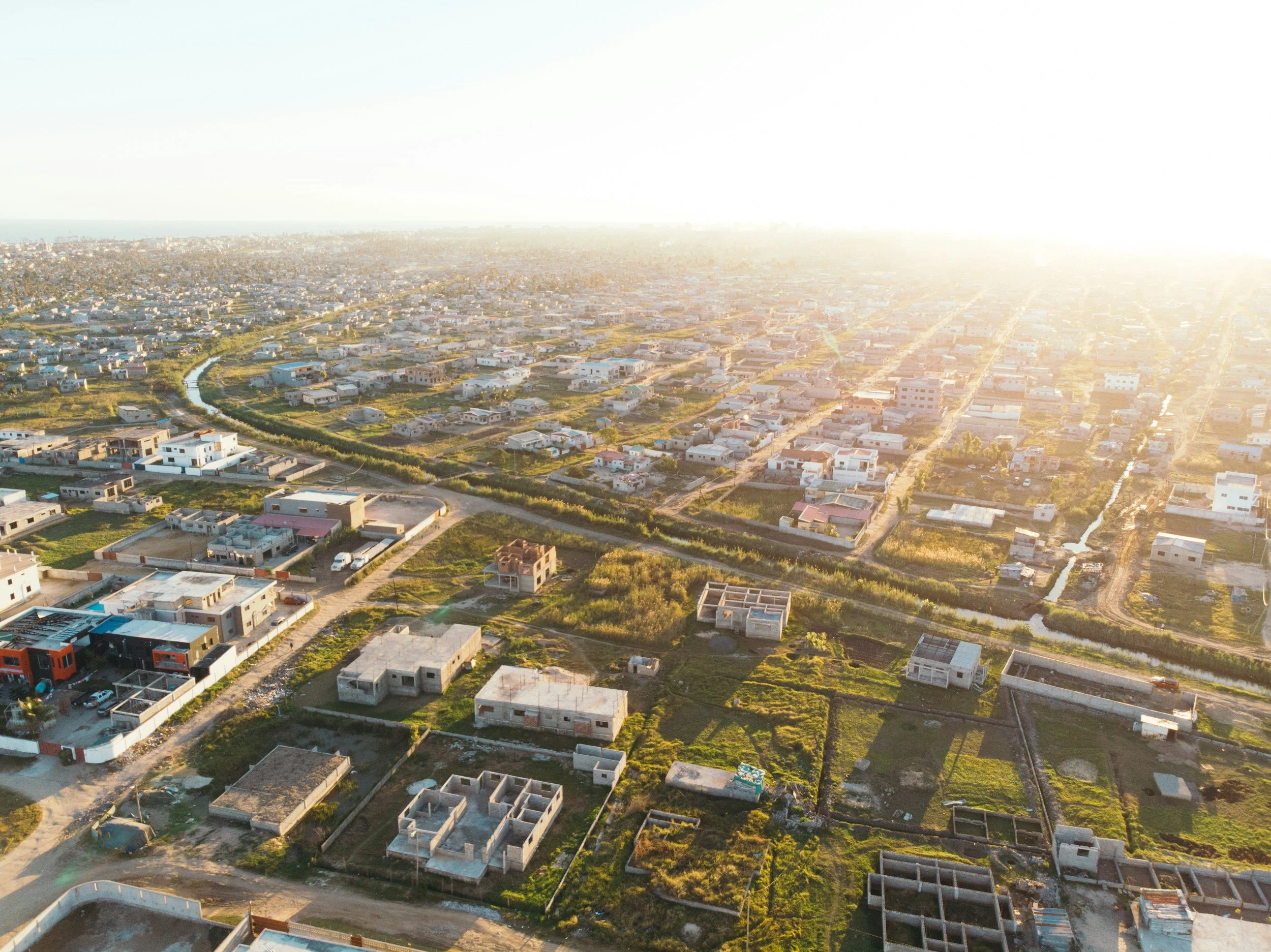 Aerial view of a suburban residential area with numerous houses and green spaces, taken during sunset with sunlight casting a glow over the area.