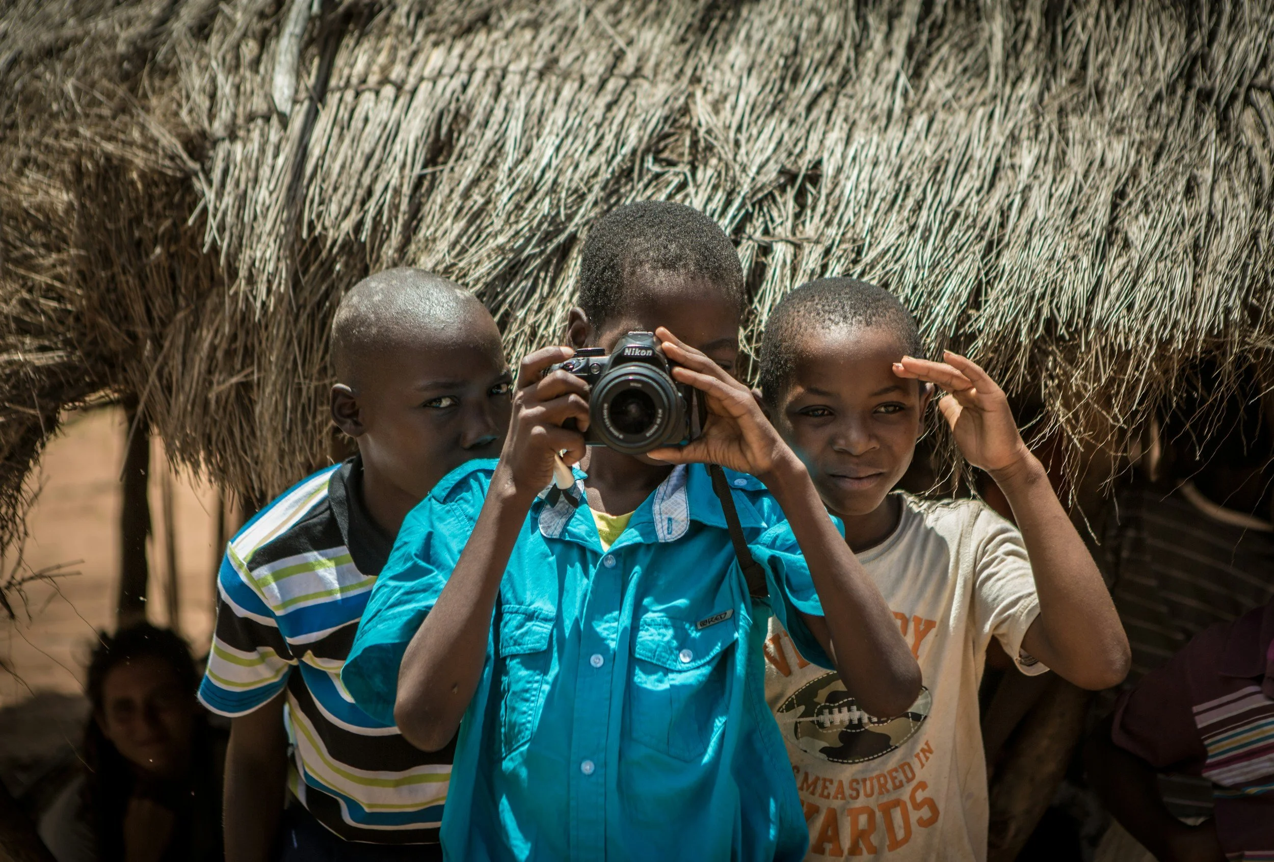 Three children under a thatched roof, with one holding a Nikon camera and taking a picture, in a rural outdoor setting.