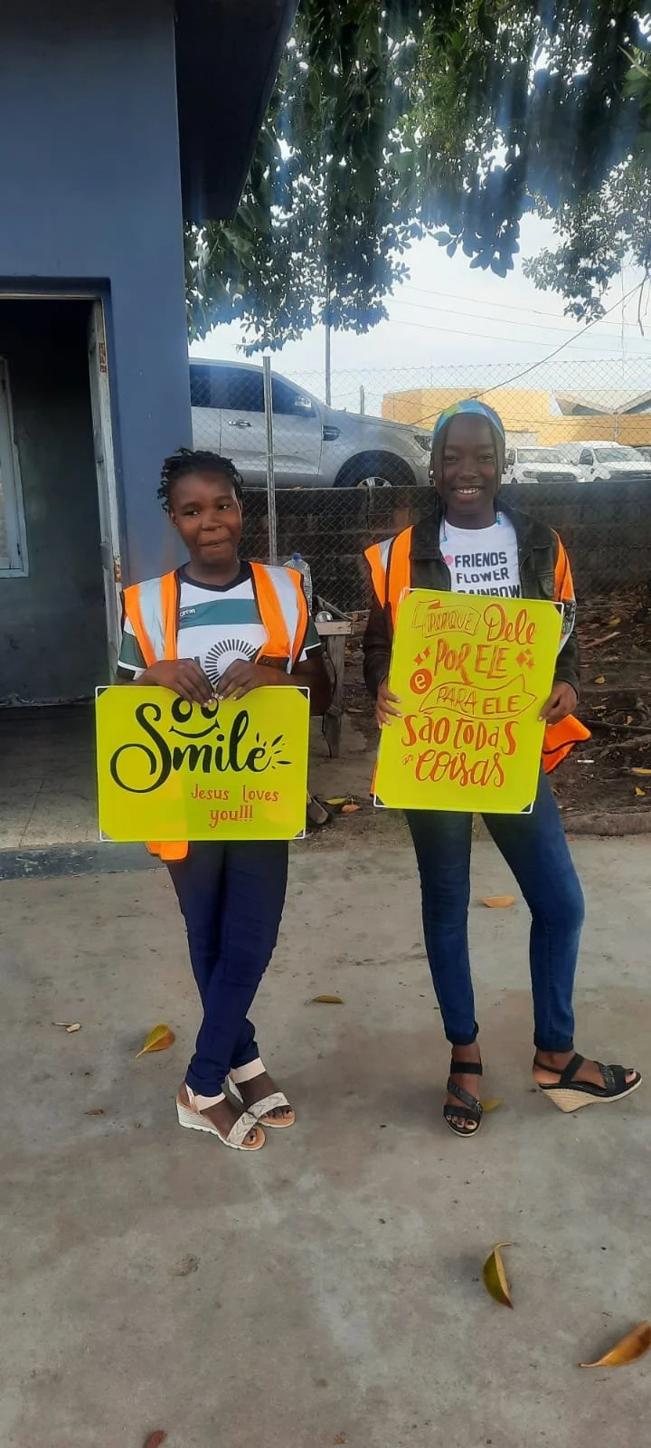 Two smiling girls holding colorful signs with messages in English and Portuguese, standing outdoors on a concrete surface with fallen leaves. The girl on the left holds a yellow sign that says "Smile" and "Jesus loves you!!!" The girl on the right holds a yellow sign with Portuguese text. Both wear orange reflective vests, casual clothes, and sandals.