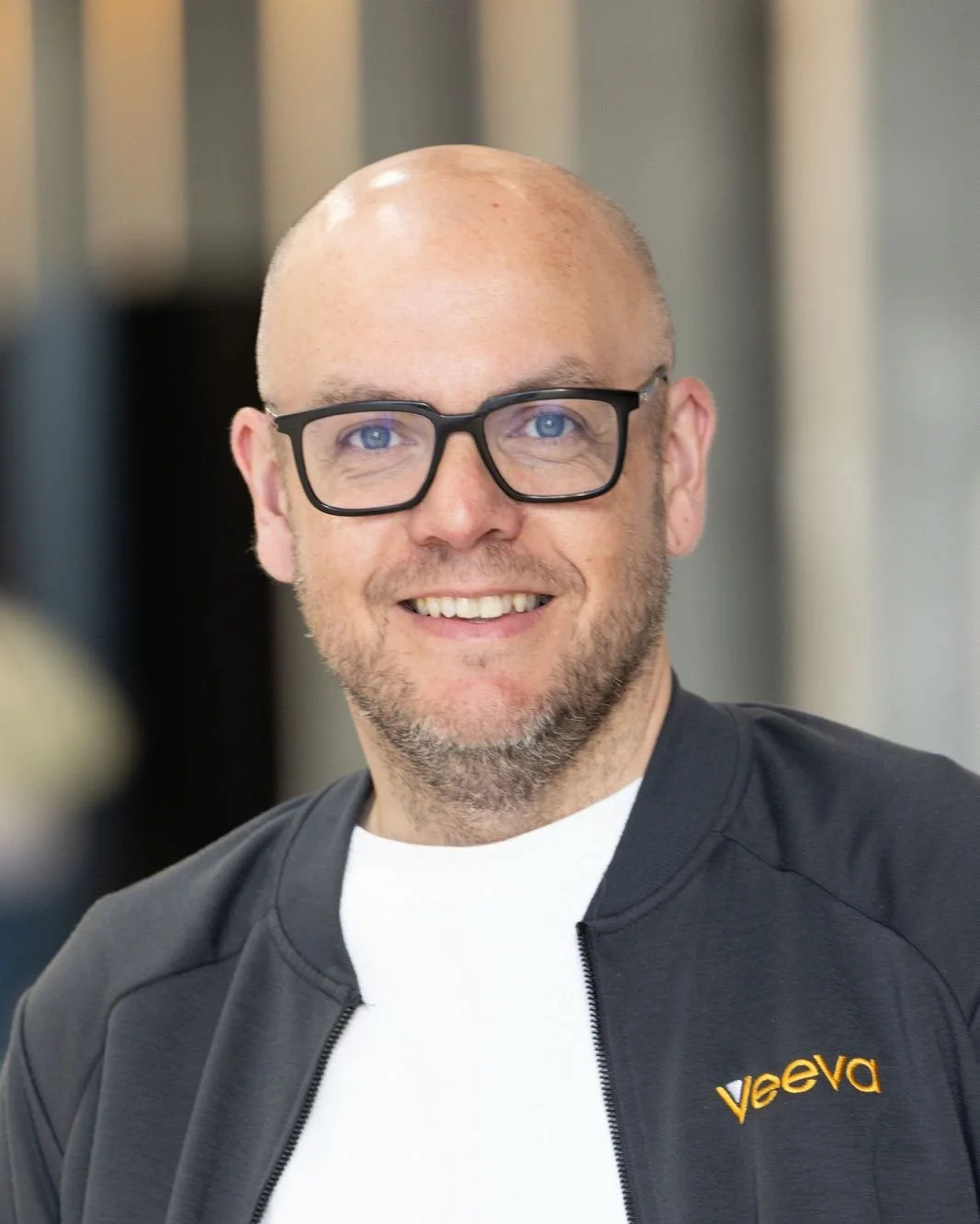 Close-up of a bald man wearing black glasses, looking into the camera, with a neutral background.