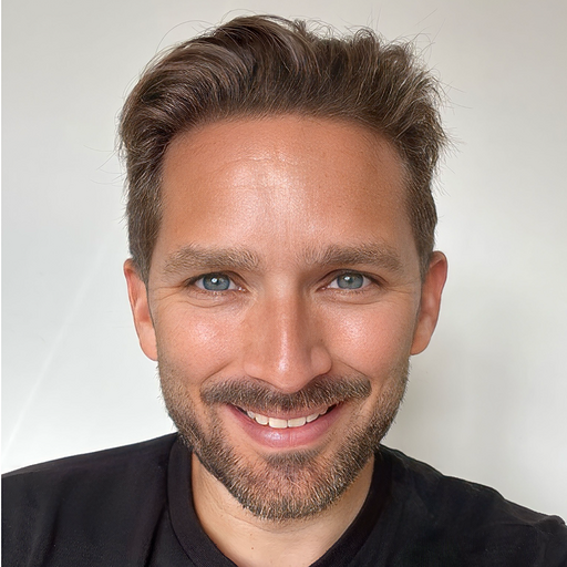 Close-up of a smiling man with short, light brown hair and blue eyes, wearing a black shirt against a plain white background.