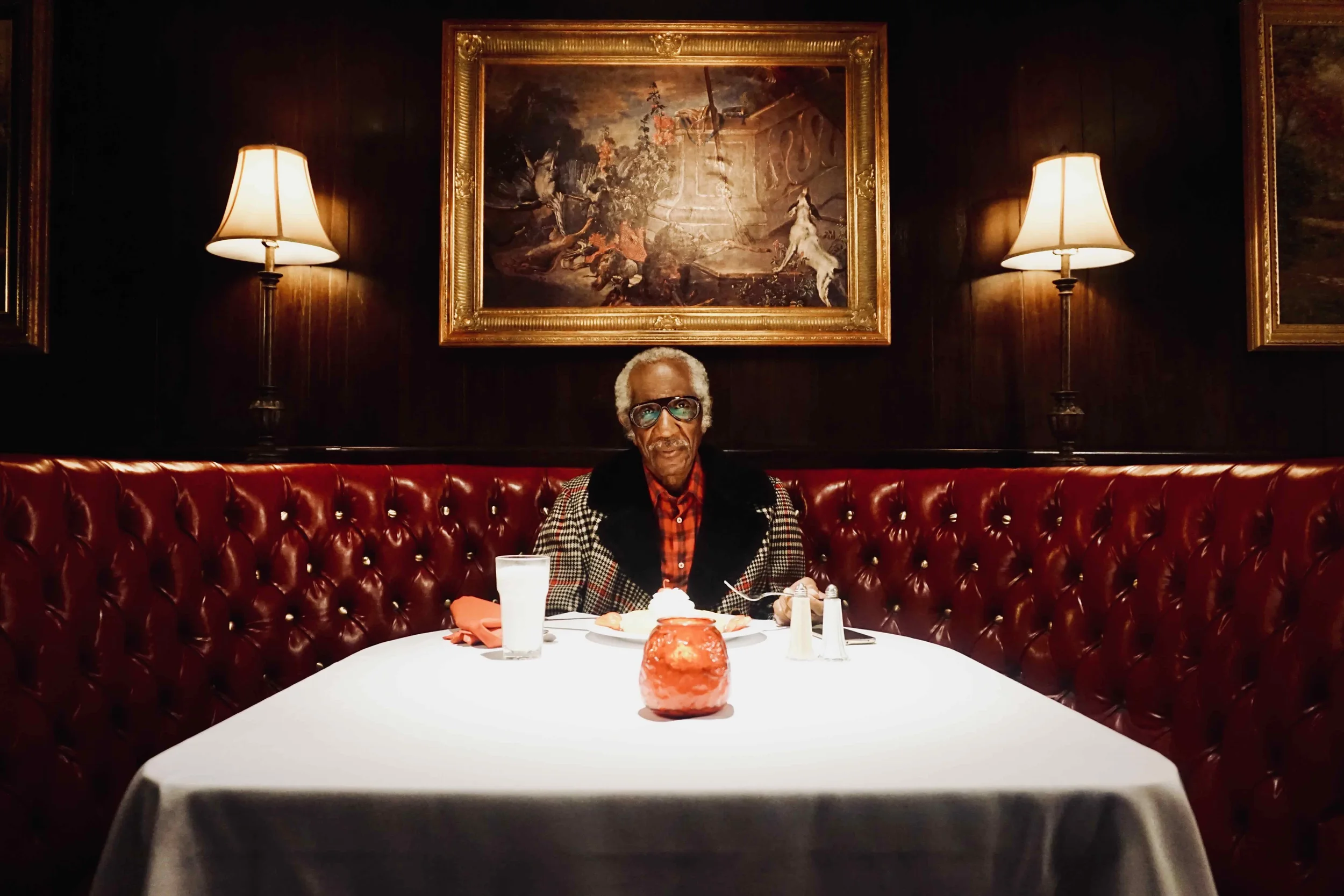 An elderly man with gray hair, glasses, and a checked shirt sits alone at a round white table in a dimly lit restaurant decorated with dark wood paneling. The table is set with a glass of milk, a candle, salt and pepper shakers, and a small plate wit