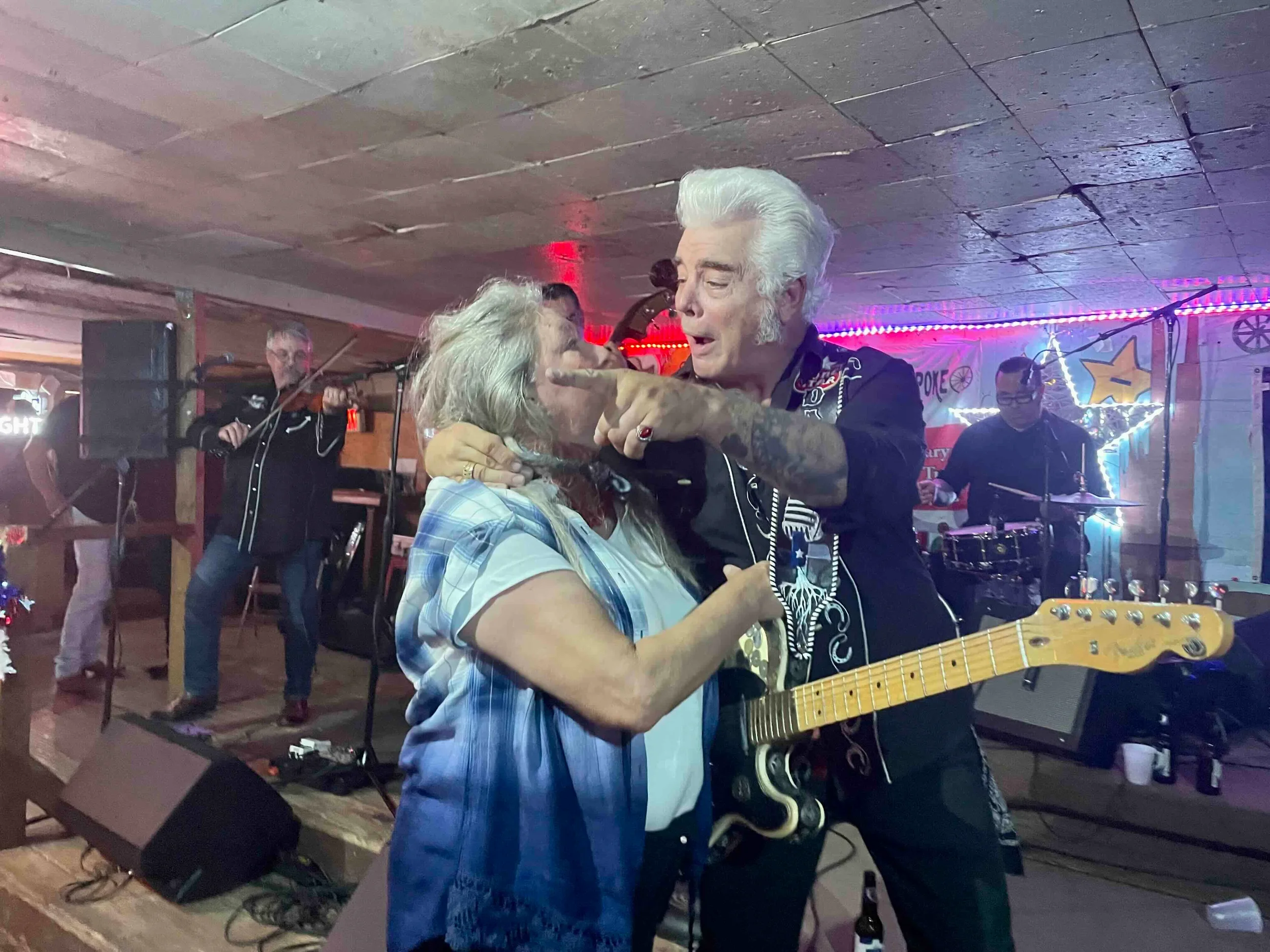 Senior man with white hair playing guitar and singing into a woman's face. The woman has gray hair and wears a blue plaid shirt. The scene is in a decorated venue with live band and colorful lights.