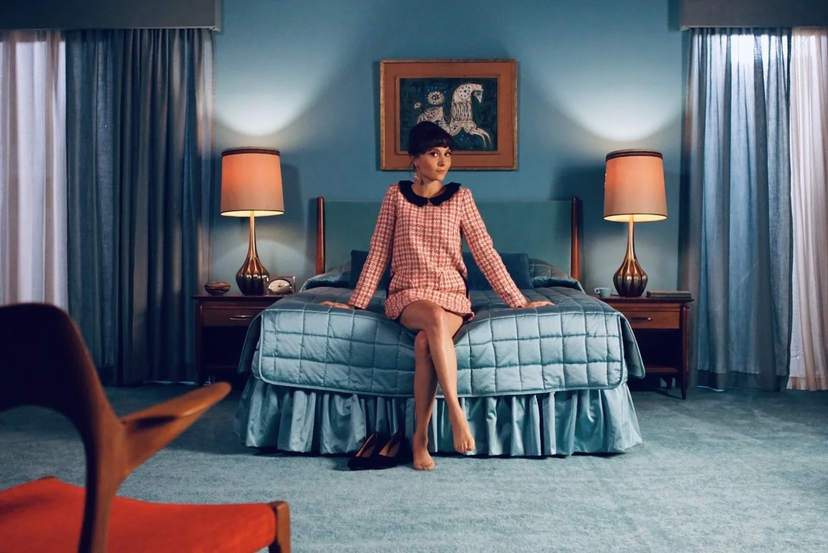 A woman sitting on a bed in a vintage-style hotel room, with her legs crossed and shoes off, looking to the side. The room has blue walls, nightstands with lamps, and curtains. There is a framed art piece of a horse hanging above the headboard.