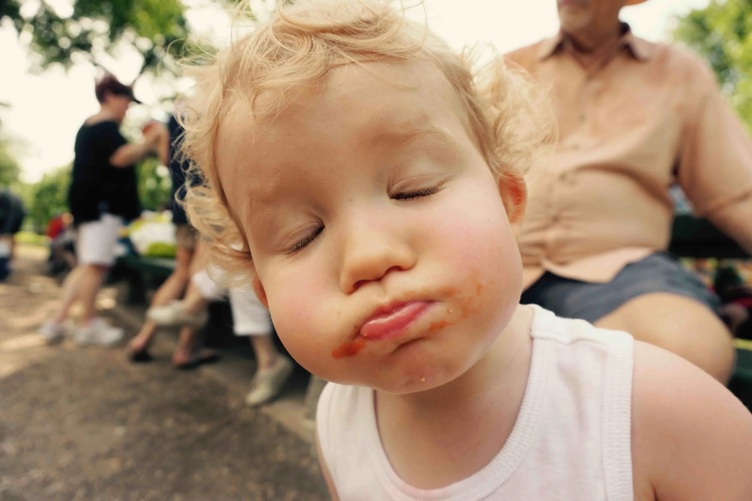 Close-up of a young child with curly blond hair and food on face, eyes closed, outdoors in a park with people in the background.