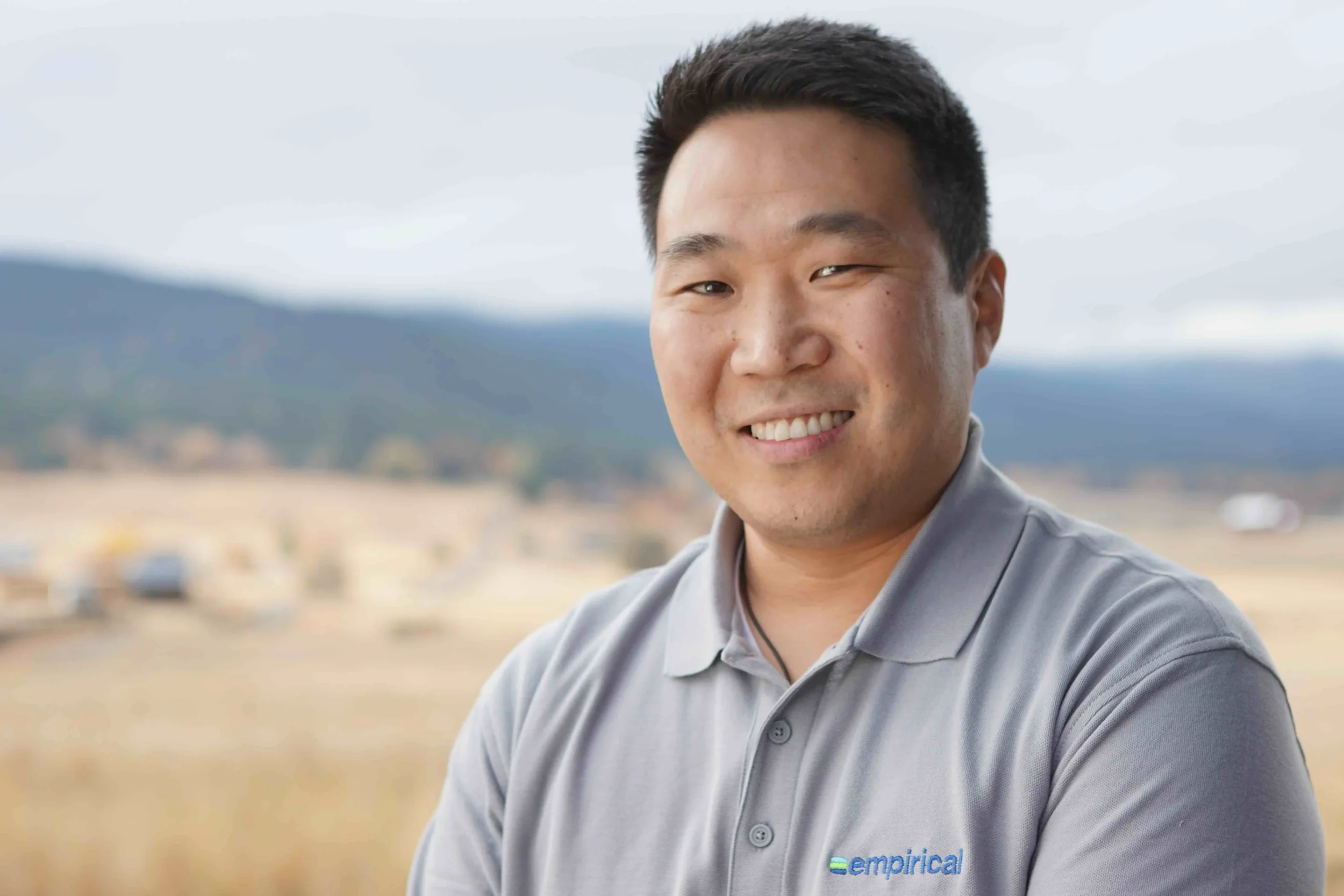 Portrait of a smiling man in a gray polo shirt standing outdoors with a blurred landscape and mountains in the background.