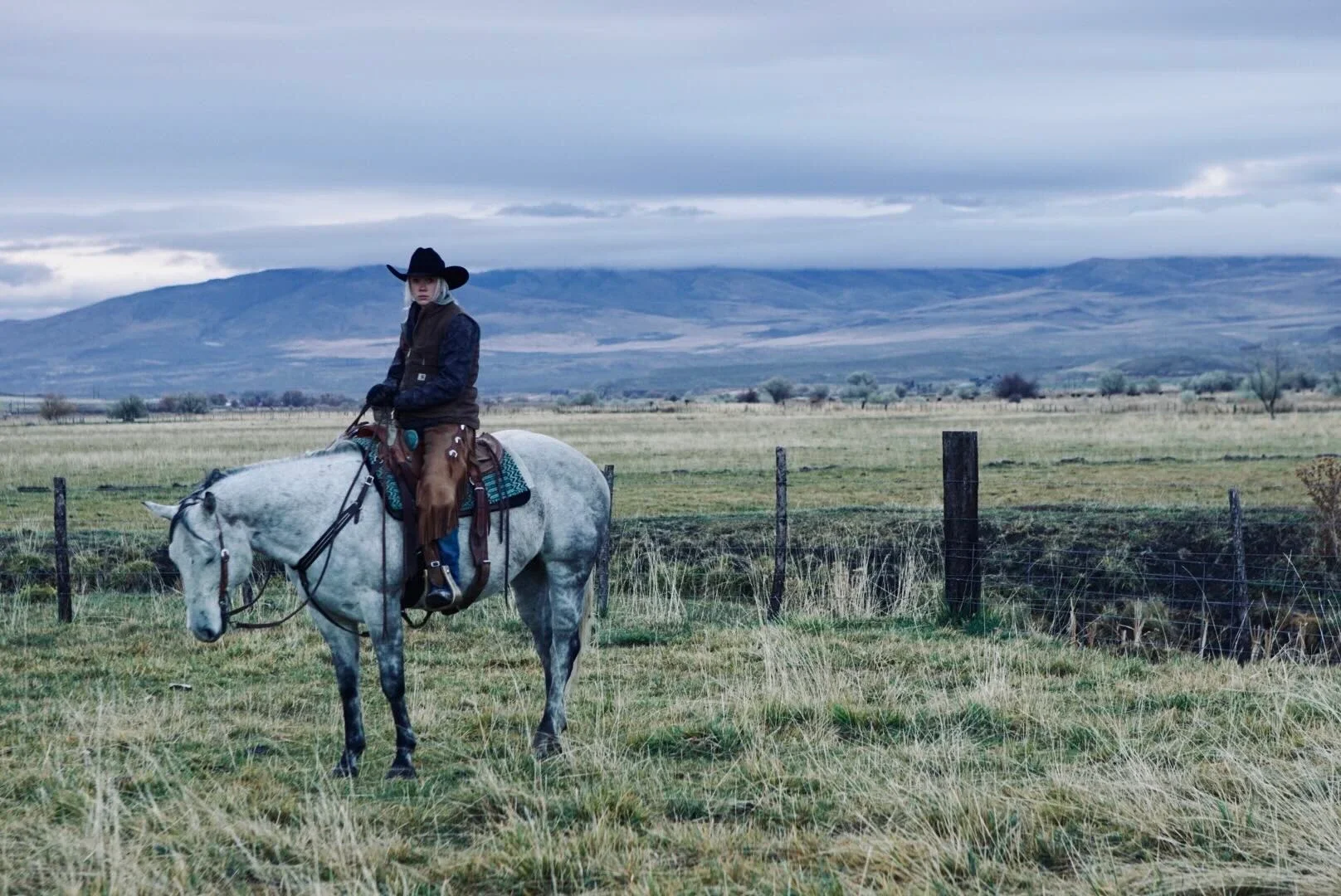 A woman in cowboy hat and dark jacket riding a gray horse in a grassy field with a fence, mountains, and cloudy sky in the background.
