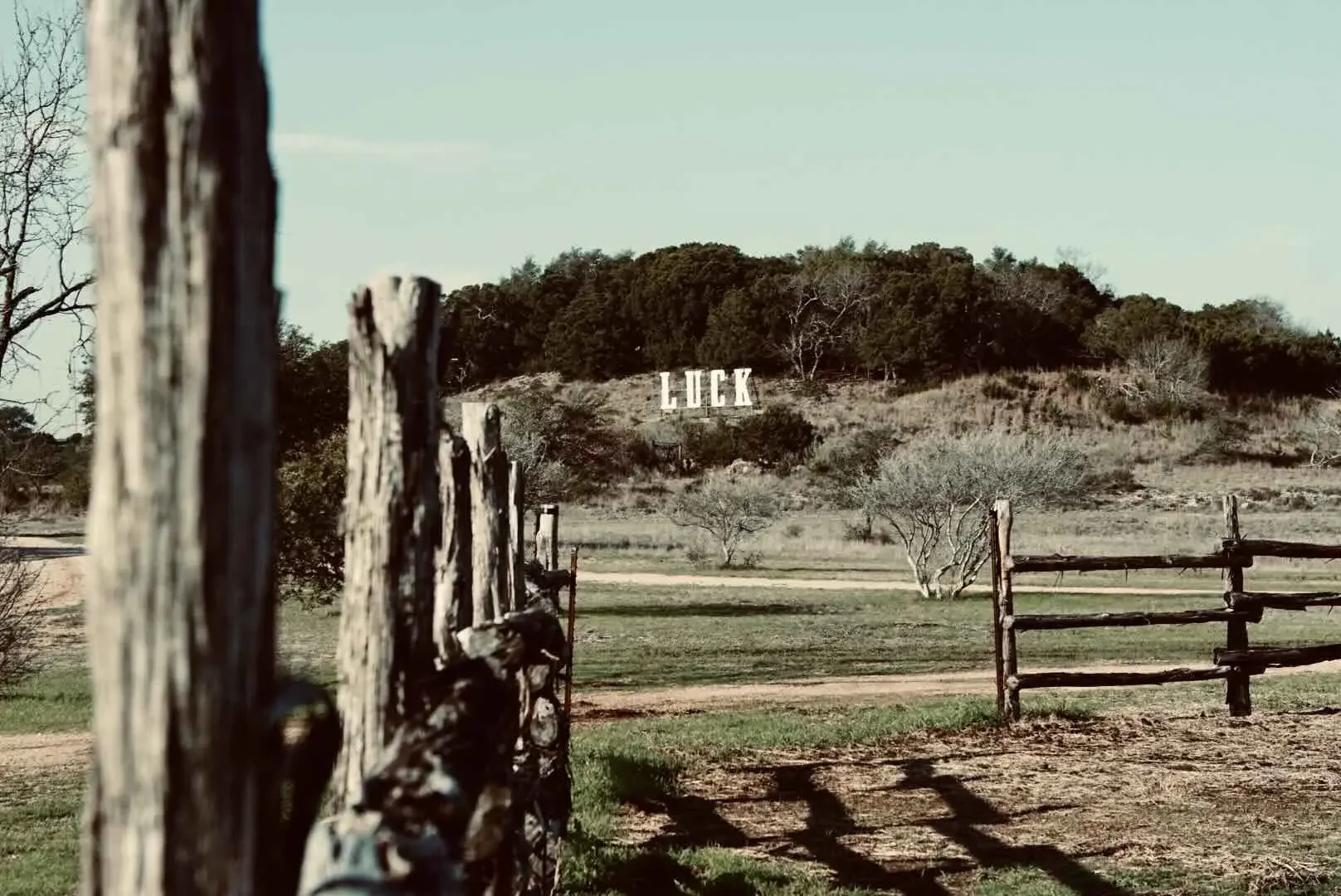A rural scene with a wooden fence, open field, and trees, with a hillside in the background displaying large white letters spelling "LUCK."
