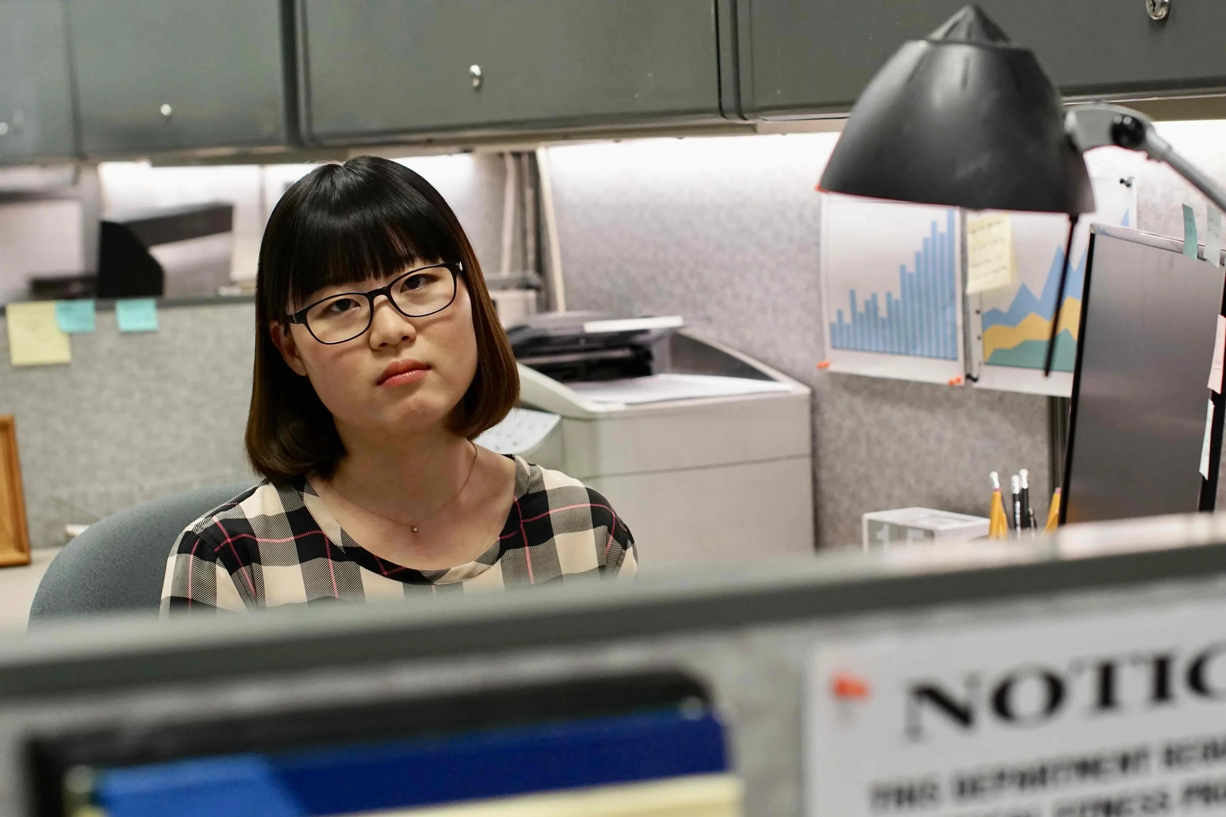 A woman with black hair, glasses, and wearing a plaid shirt sitting at a desk in an office environment, looking towards the camera with a neutral expression.
