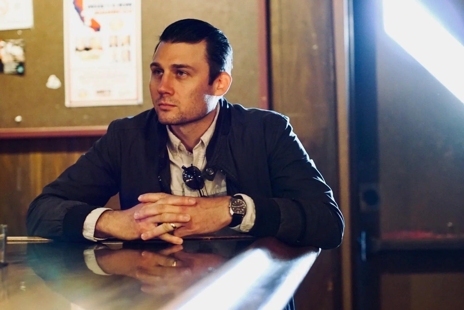 A man with dark hair, wearing a black jacket, sitting at a bar or counter with his hands clasped, looking contemplative.