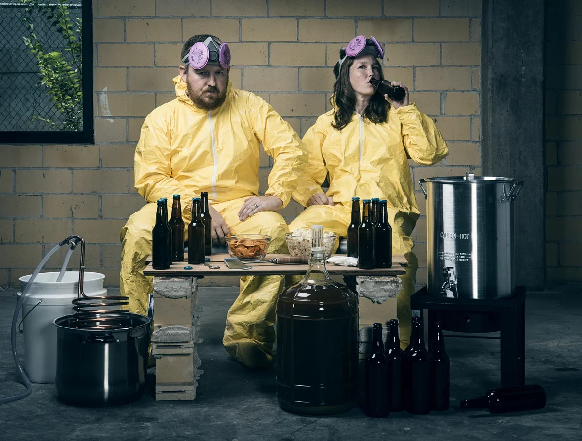 Two people in yellow hazmat suits sitting on a bench, surrounded by beer bottles and brewing equipment, with one drinking from a bottle and the other with a serious expression.