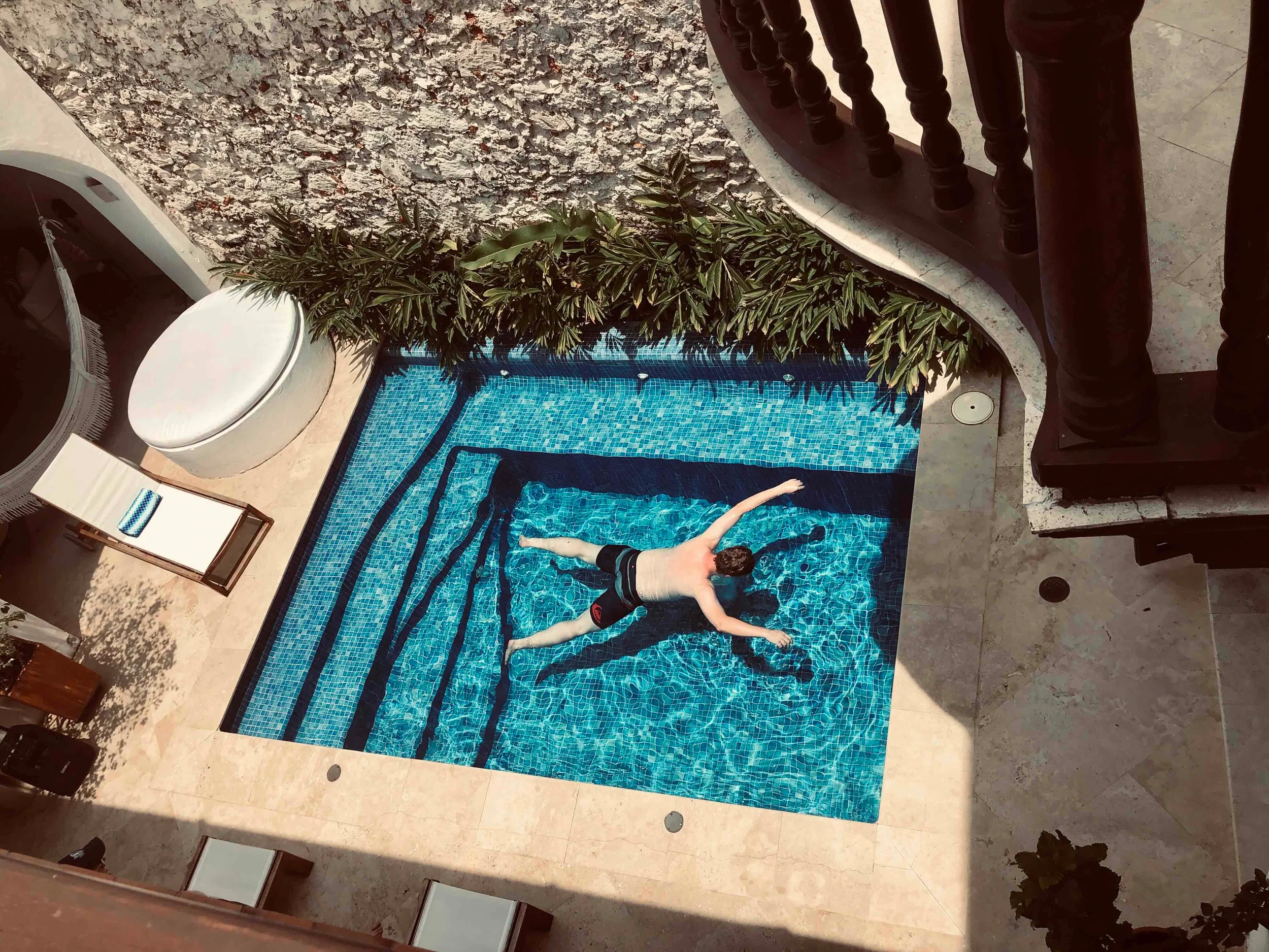 A person is floating on their back in a small rectangular swimming pool with blue tile interior, surrounded by a stone patio and a wall with greenery.