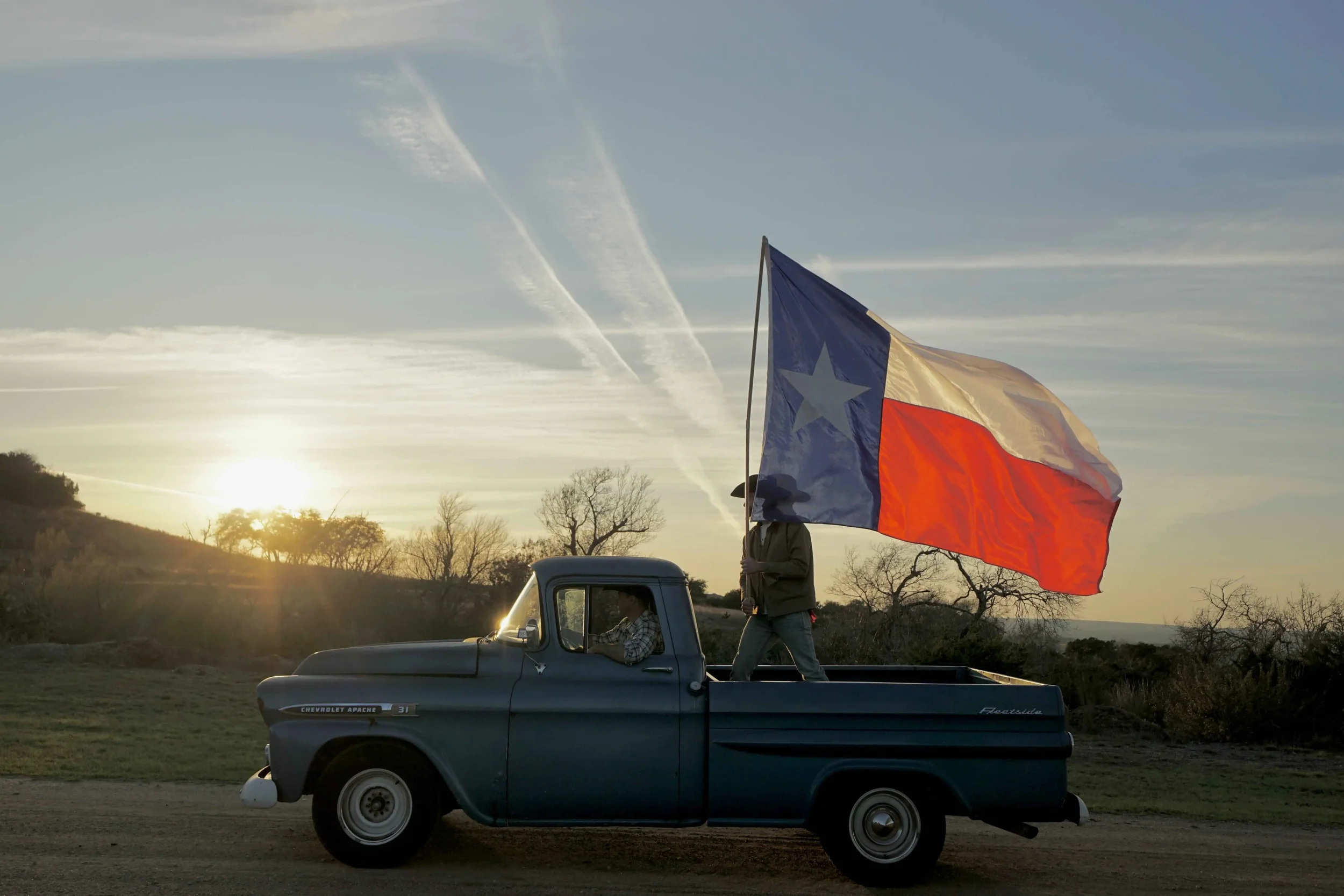 A vintage Chevrolet Apache pickup truck parked on a dirt road at sunset with a person standing in the truck bed holding a large Texas flag. The scene has a rural landscape with trees and hills in the background.