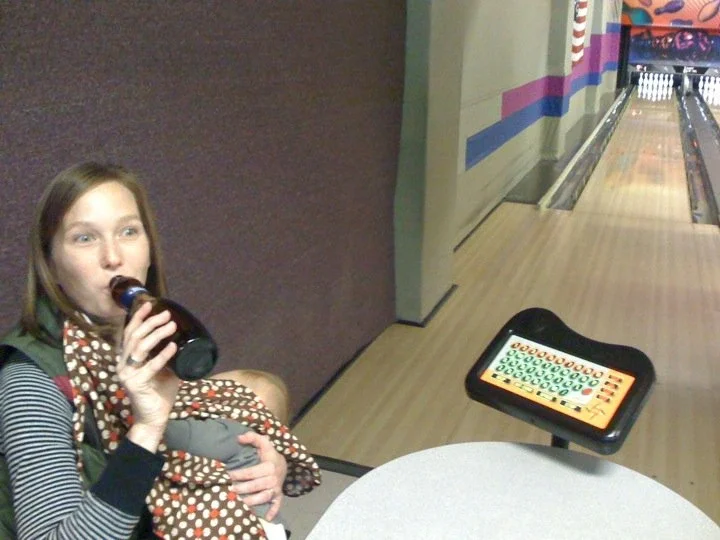 A young girl sitting at a table in a bowling alley, drinking a soda. She has a baby wrapped in a polka-dot blanket on her lap, and a bowling lane with pins in the background.