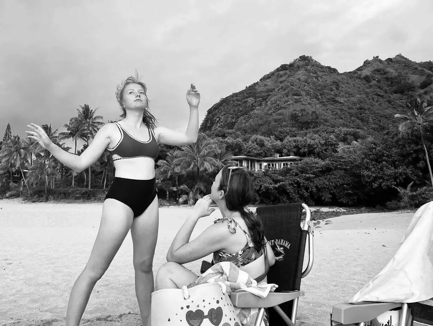 A woman in a black swimsuit is standing on a beach, striking a pose, while another woman sits nearby in a foldable chair, listening attentively. The background features lush tropical vegetation and hills.