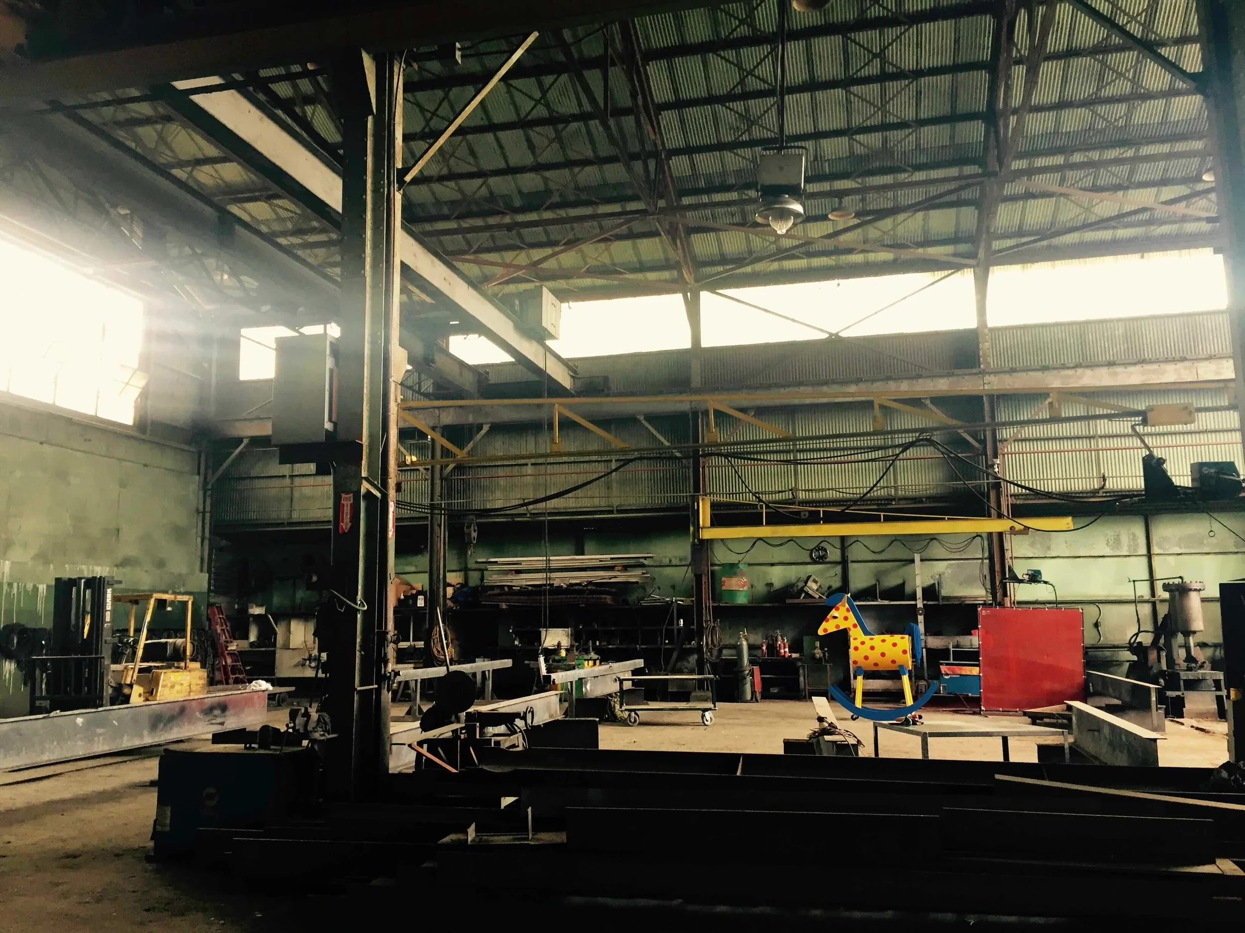 Interior of a warehouse or workshop with metal beams and a corrugated metal roof. There are various tools and equipment scattered throughout, including a colorful rocking horse on the right side.