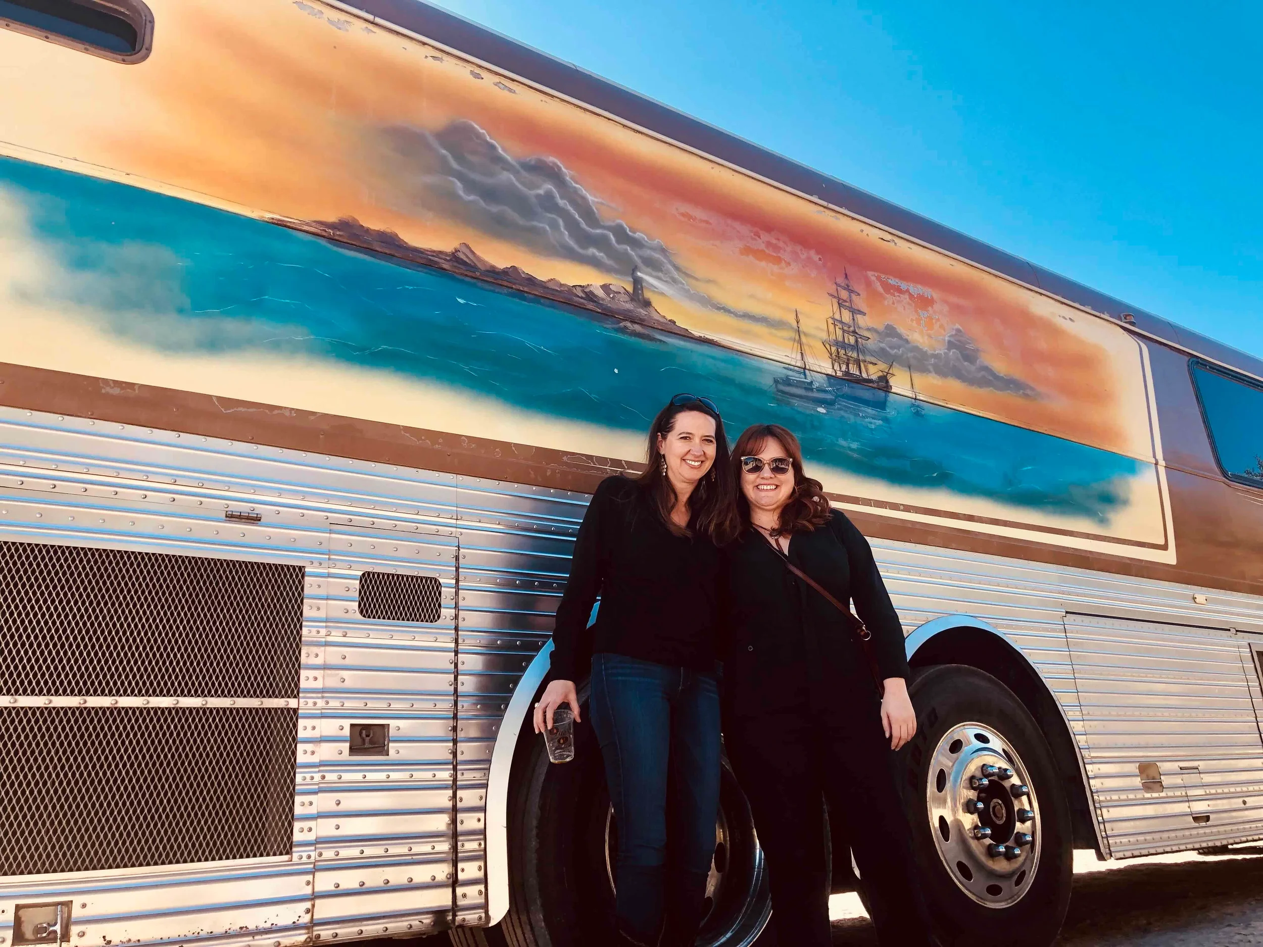 Two women stand in front of a painted bus featuring a sunset, ocean waves, mountains, and ships.