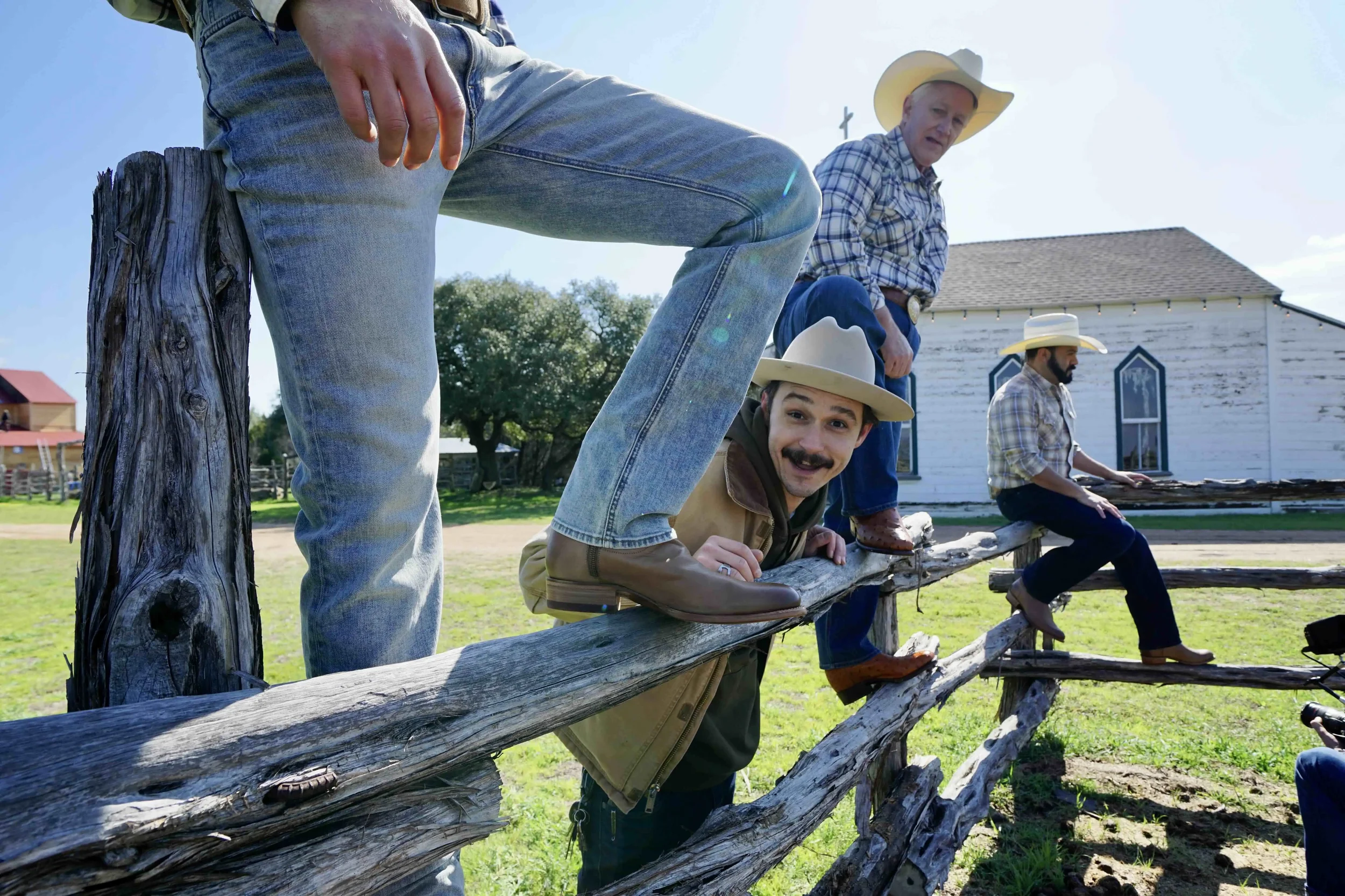 People dressed as cowboys on a wooden fence in a rural setting with a white barn and trees, sunny sky.