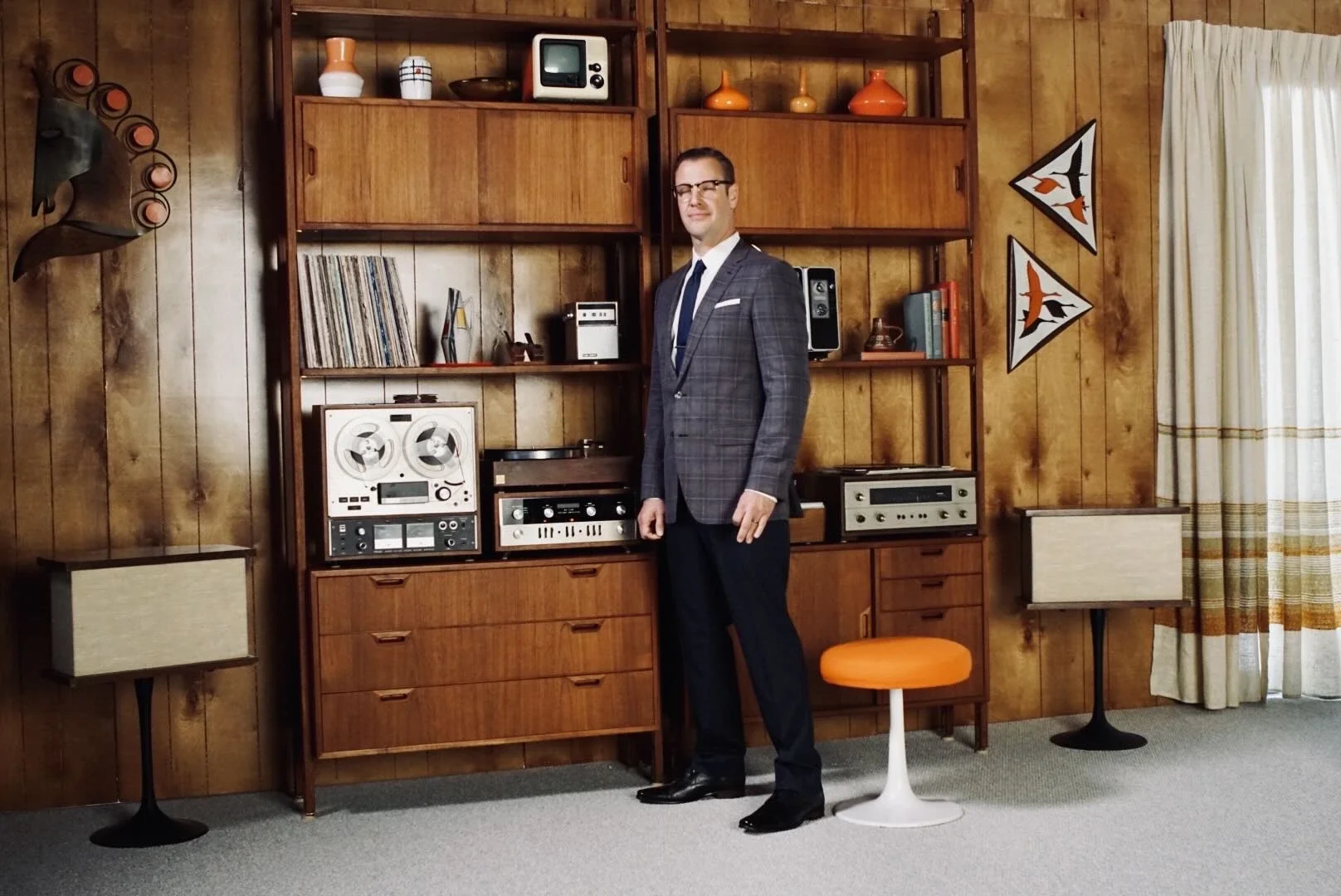 A man in a plaid suit and glasses standing in a vintage living room with wooden wall paneling, surrounded by retro decor including vinyl records, stereo equipment, and patterned curtains.