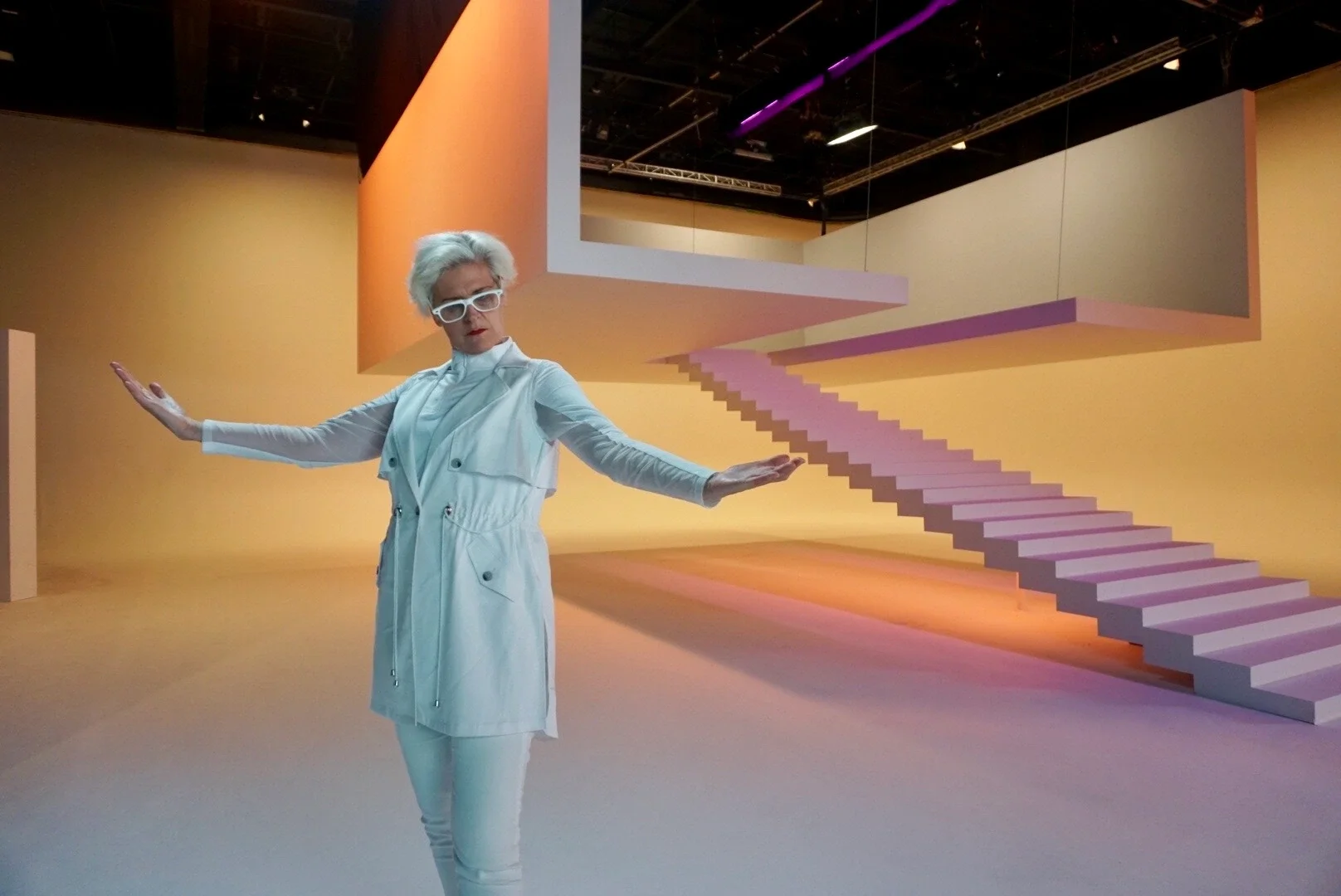 An elderly woman with white hair, wearing white glasses and a white outfit, standing with arms outstretched in an art installation featuring a large pink staircase and geometric shapes in a gallery setting with colorful lighting.
