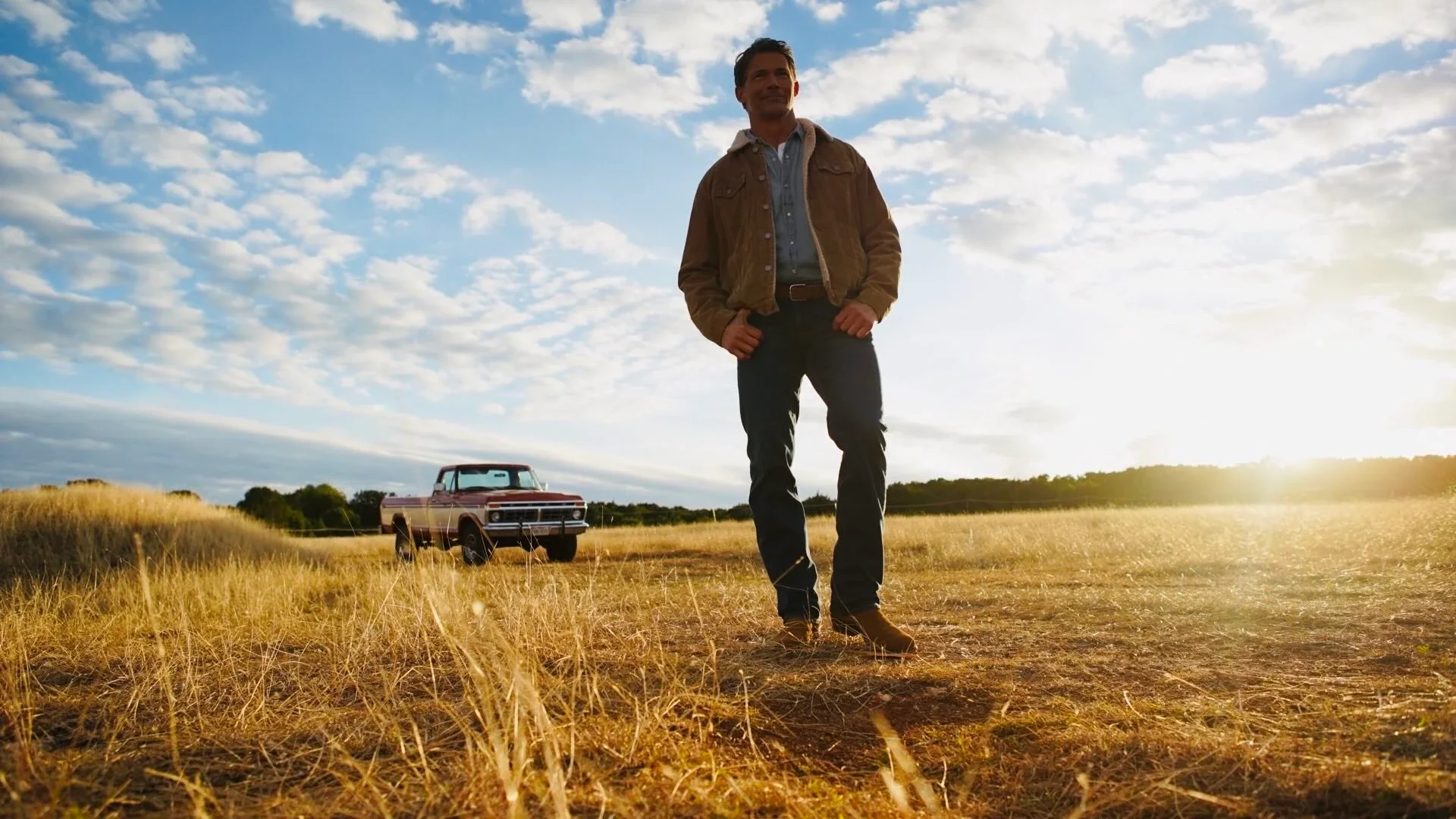 A man standing in a grassy field during sunset, with a vintage truck in the background.