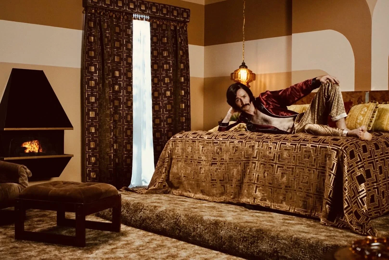 A man lounging on a bed in a richly decorated bedroom with patterned curtains and vintage-style lighting.