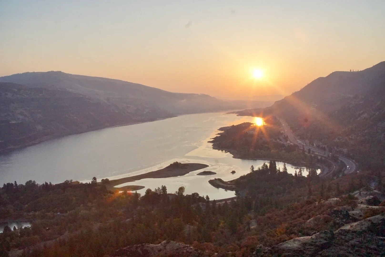 A river flowing through a valley with mountains on either side during sunset, with the sun partially behind the mountains reflecting on the water.