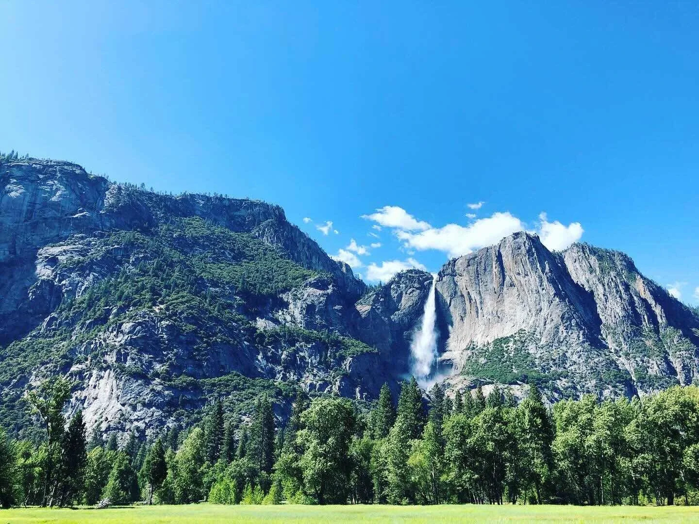 Scenic view of mountains with a waterfall, lush green trees in the foreground, and a bright blue sky with some clouds.